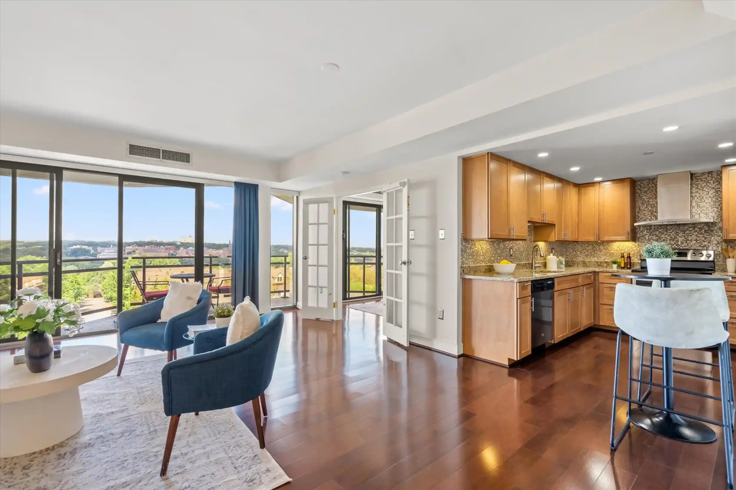 A modern living room and kitchen with large sliding glass doors opening to a balcony, with city and green landscape views