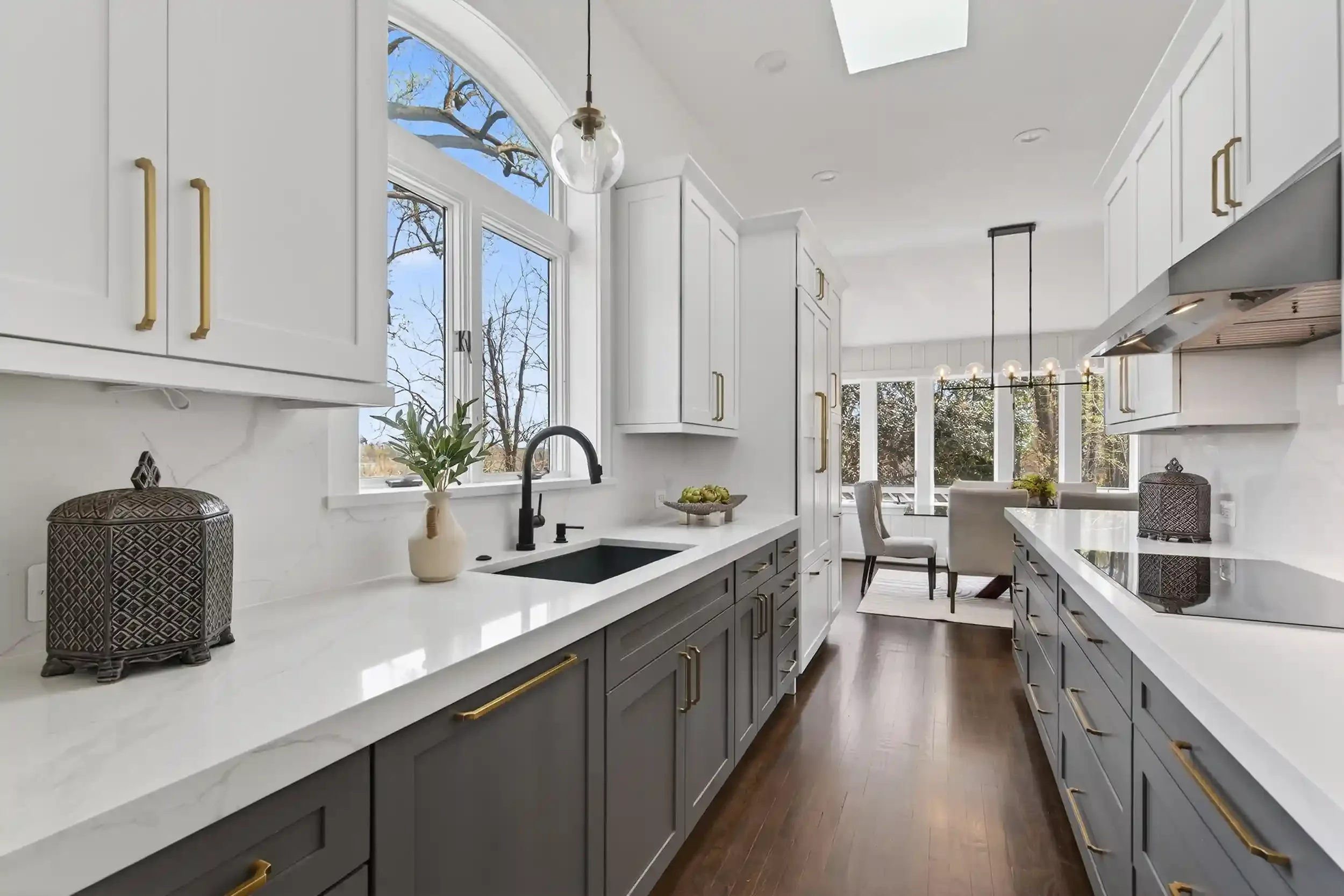 Modern kitchen with white backsplash, gray lower cabinets with gold handles, white countertops, and large windows letting in natural light. Decor includes a decorative jar, a plant, and a fruit bowl