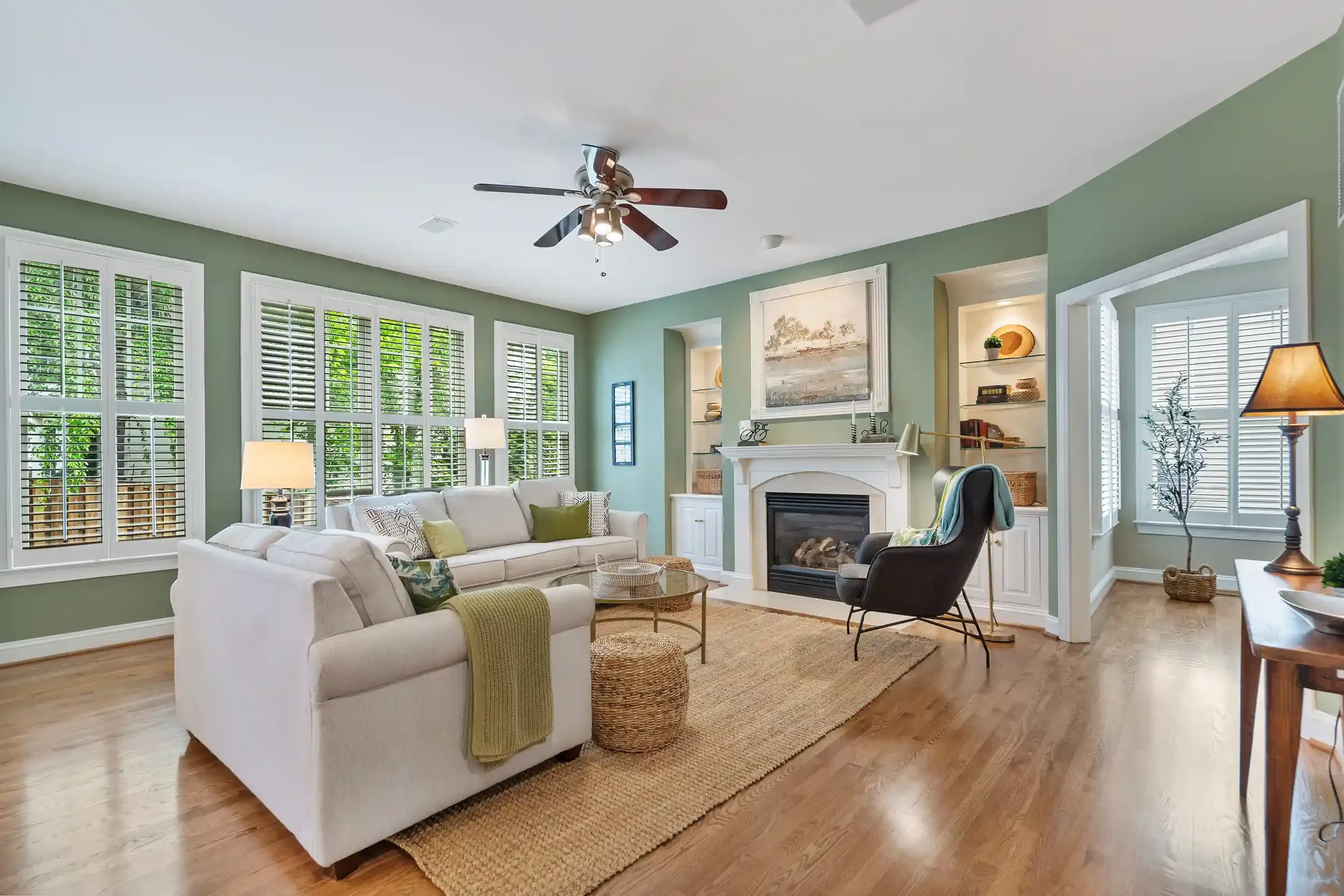 A living room with green walls, large windows with white shutters, a white sofa with patterned pillows, and a black armchair
