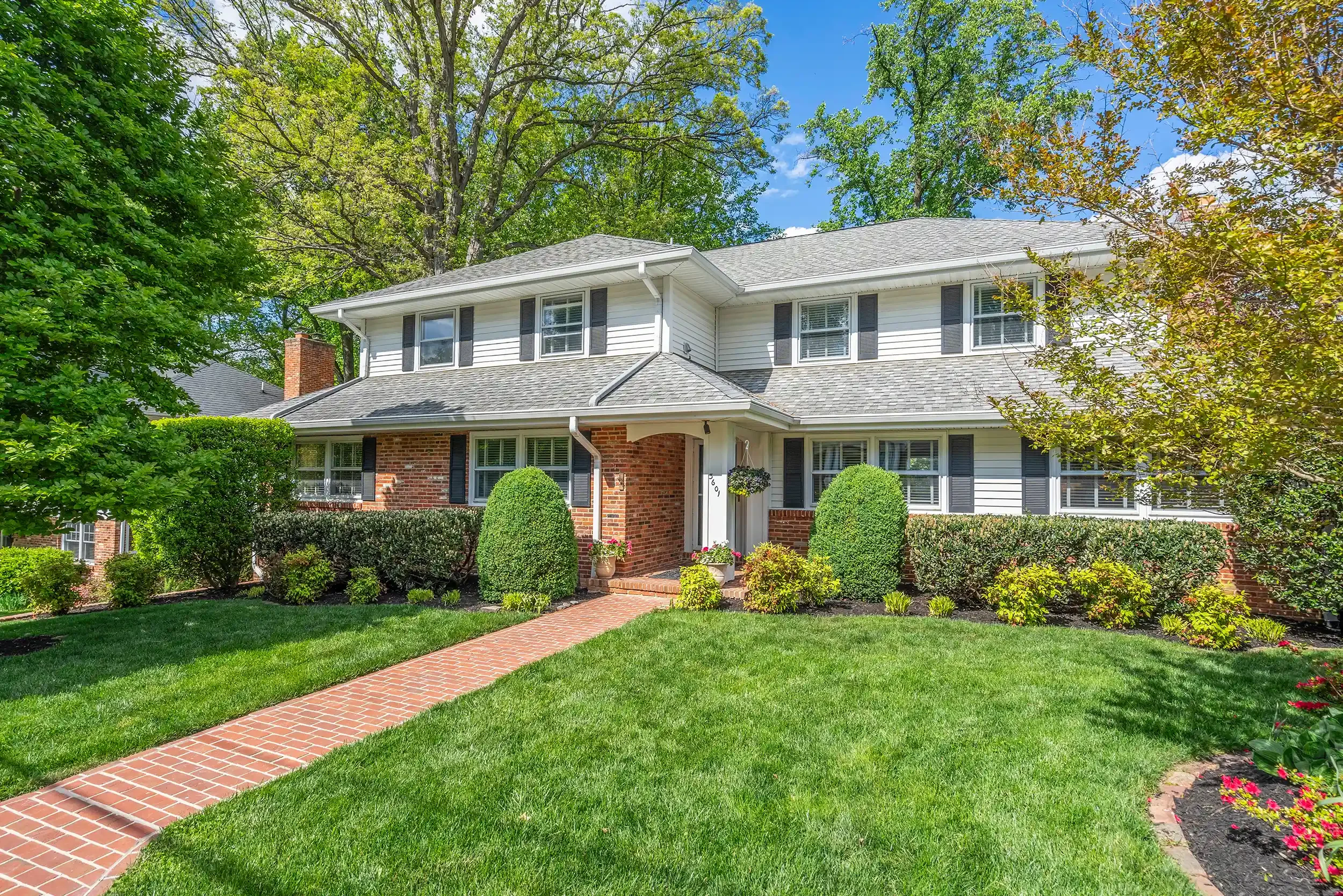 A white two-story house with black shutters, brick accents, and a gable roof, surrounded by green trees and a well-maintained lawn with a brick pathway leading to the front door