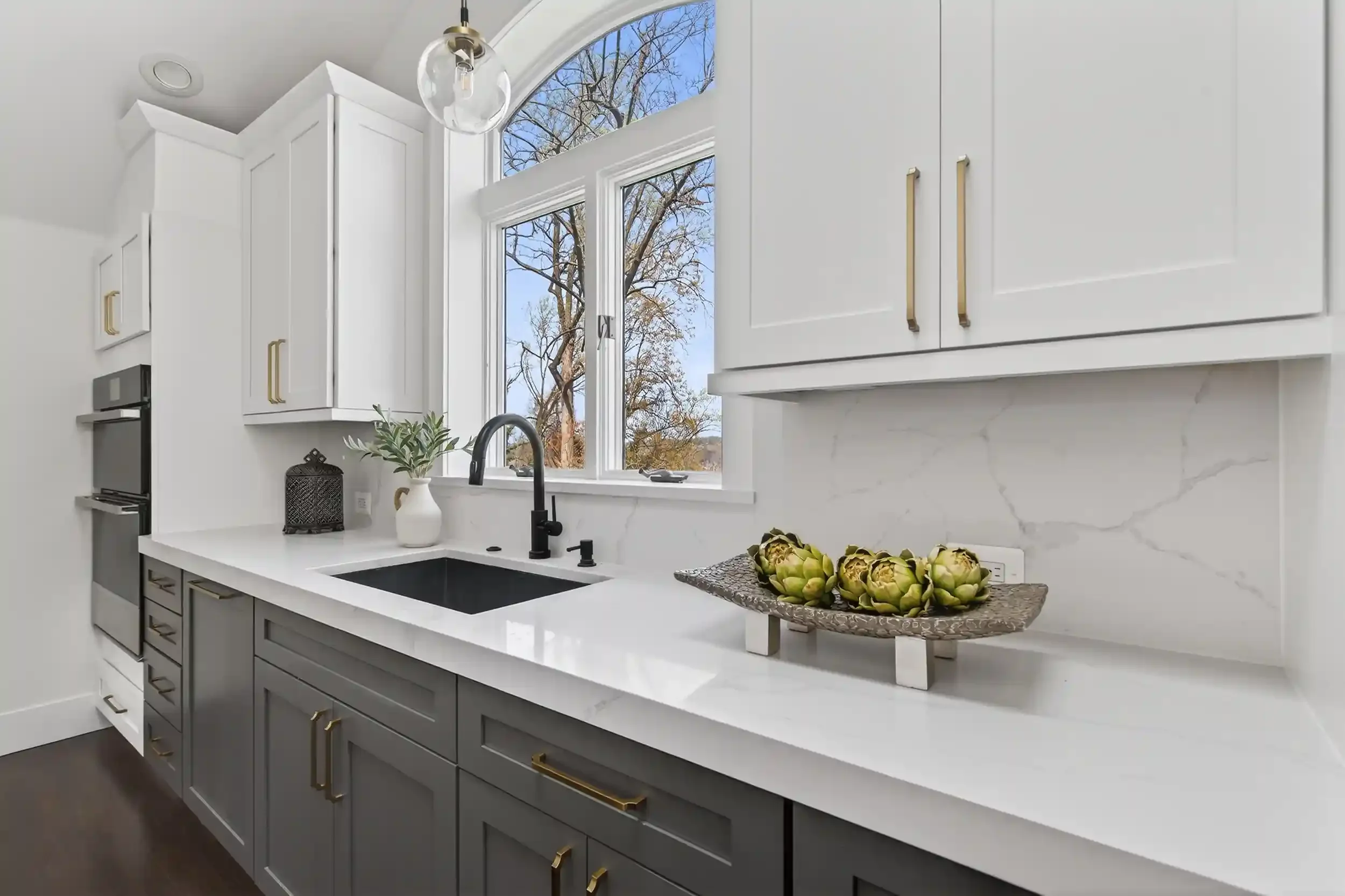 A modern kitchen with white upper cabinets and gray lower cabinets, a white countertop with a black sink, gold handles, a decorative bowl of artichokes, a potted plant, and large windows