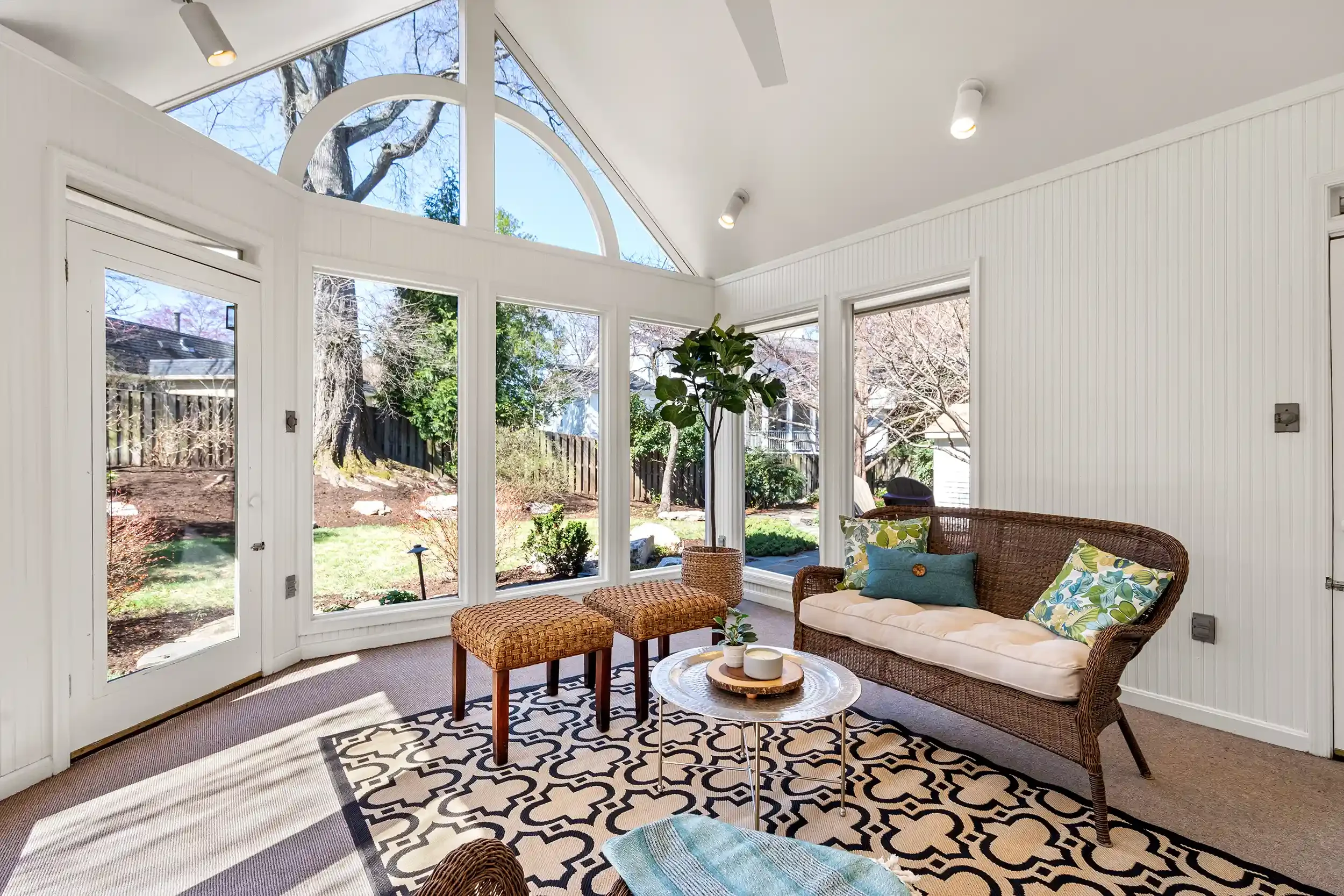 Sunroom with large windows, wicker furniture with floral and solid cushions, a round coffee table with decorative items, patterned area rugs, and a view of the backyard with trees and a fence