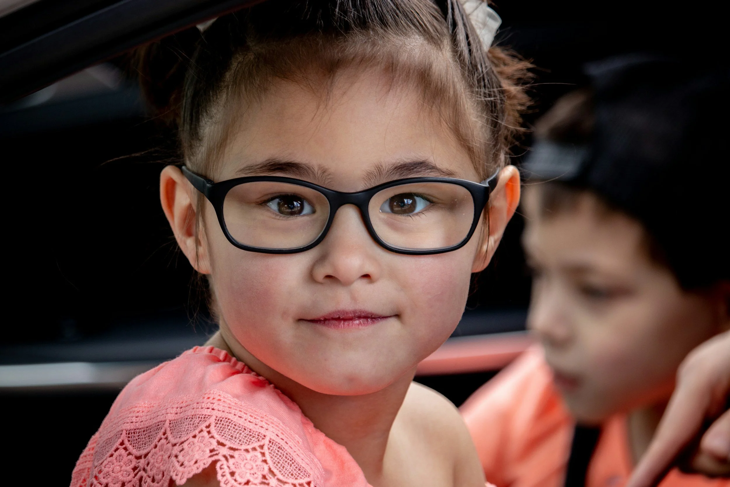 Close-up of a young girl with glasses and brown hair, wearing a pink shirt, looking at the camera.