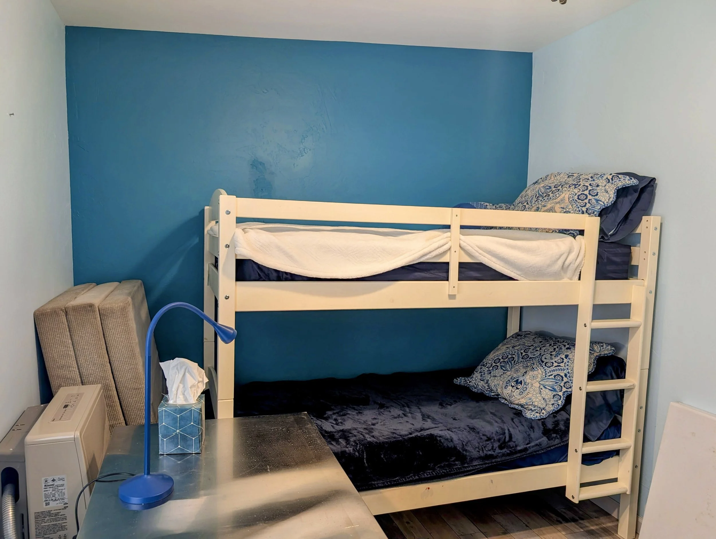 A bunk bed in a small room with a blue accent wall. The top bunk has a pillow with blue, beige, and white patterns, with a white blanket. The bottom bunk has a dark blanket and a patterned pillow that matches the top. Next to the bed, there is a smal