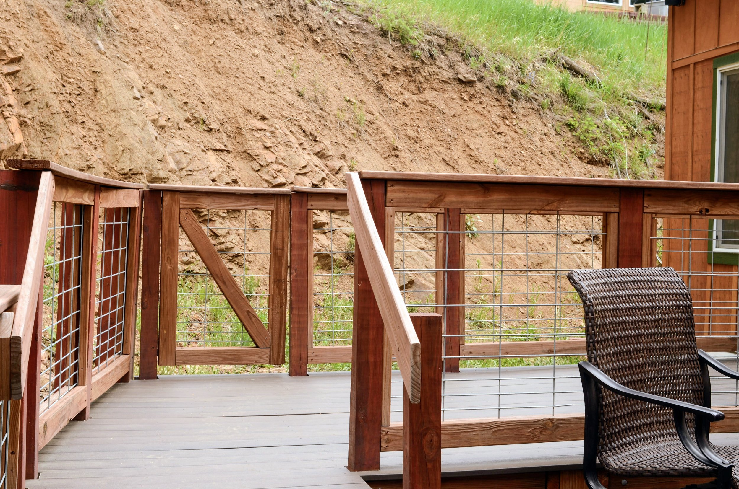 A backyard deck with a wooden railing, overlooking a dirt hillside and some grass, with part of a house visible on the right.