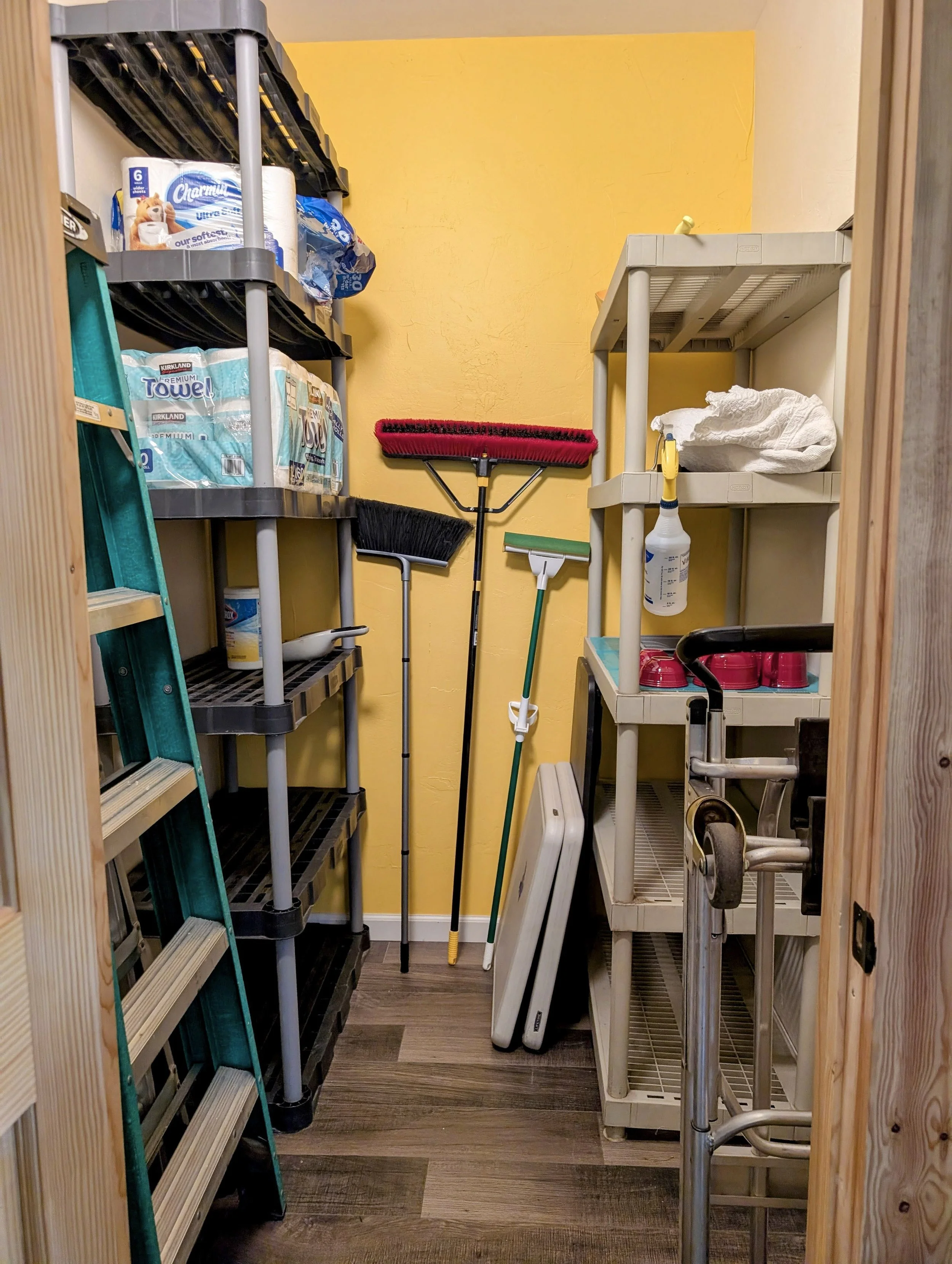 A small storage closet with metal shelves holding paper towels, cleaning supplies, and a box of cups. The walls are painted yellow, and the floor has a wood look. There are cleaning tools including a broom, dustpan, and scrub brush. A red lint roller