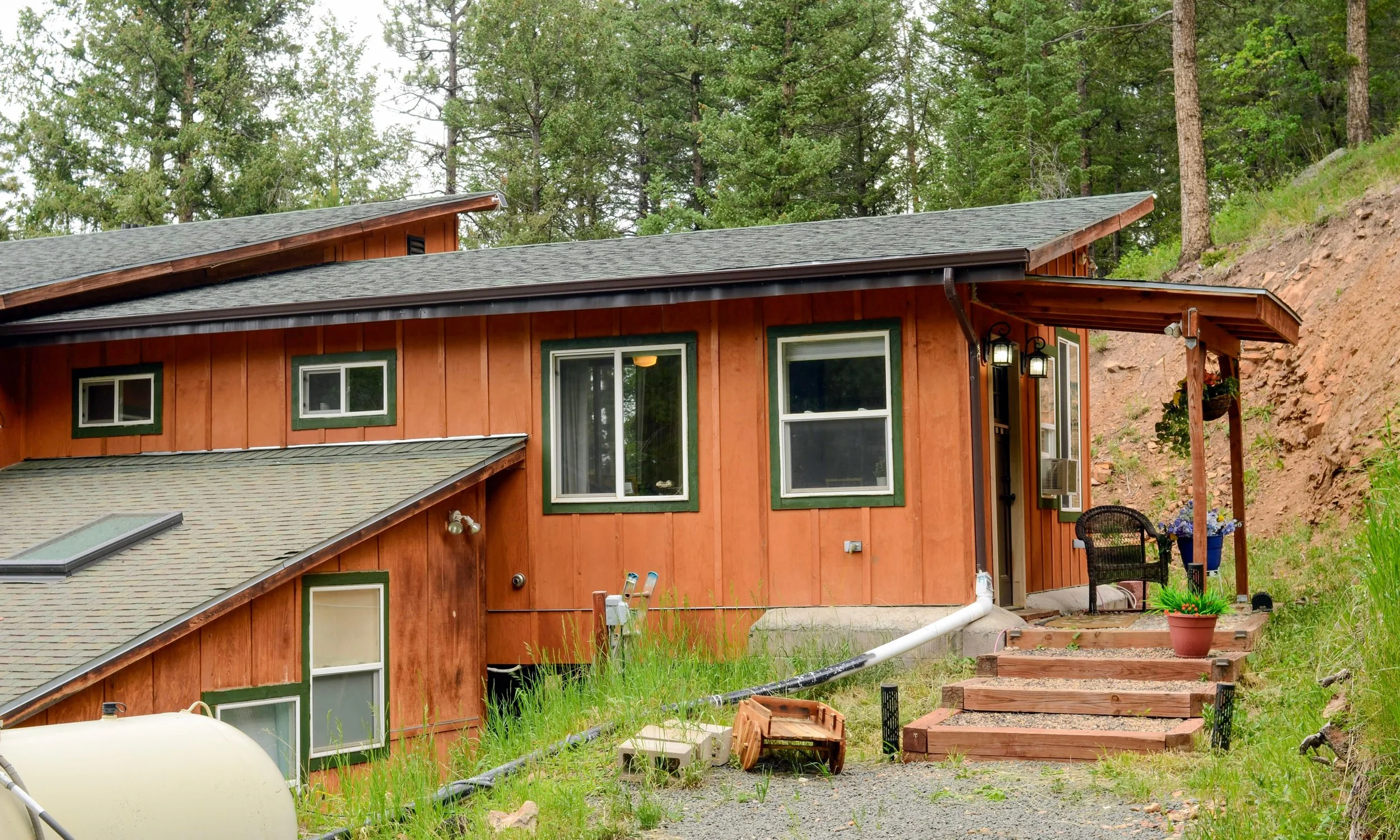 Back of a two-story wooden house with green trim, surrounded by tall trees and a grassy yard. There are steps leading up to a small porch with hanging flower pots, outdoor seating, and a gardening area in the yard.