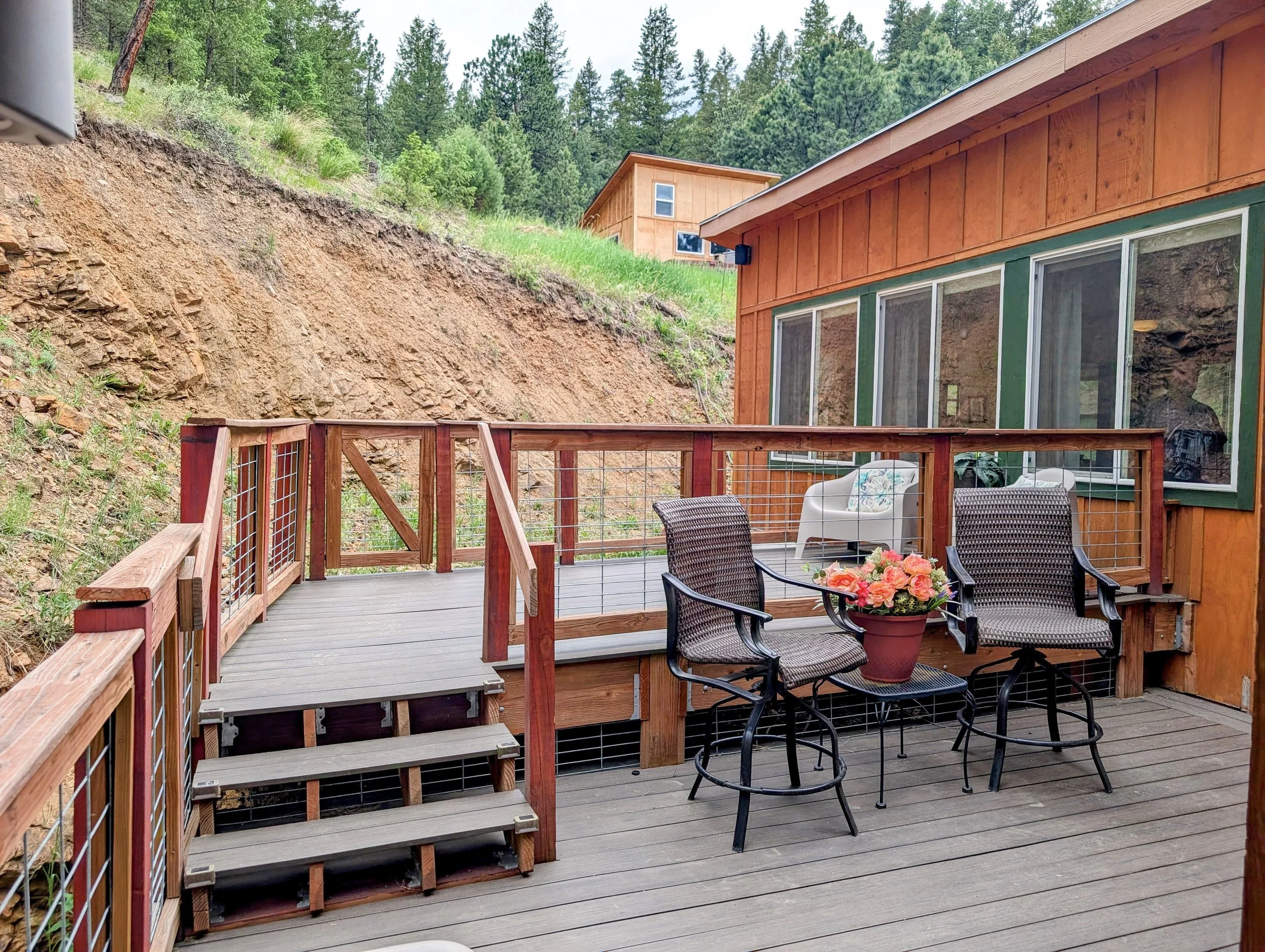 A wooden deck with outdoor furniture, including chairs and a table with flowers. A railing surrounds the deck, and a hillside with dirt and rock is visible behind it. In the background, there are green trees and a wooden house or shed on a hill.