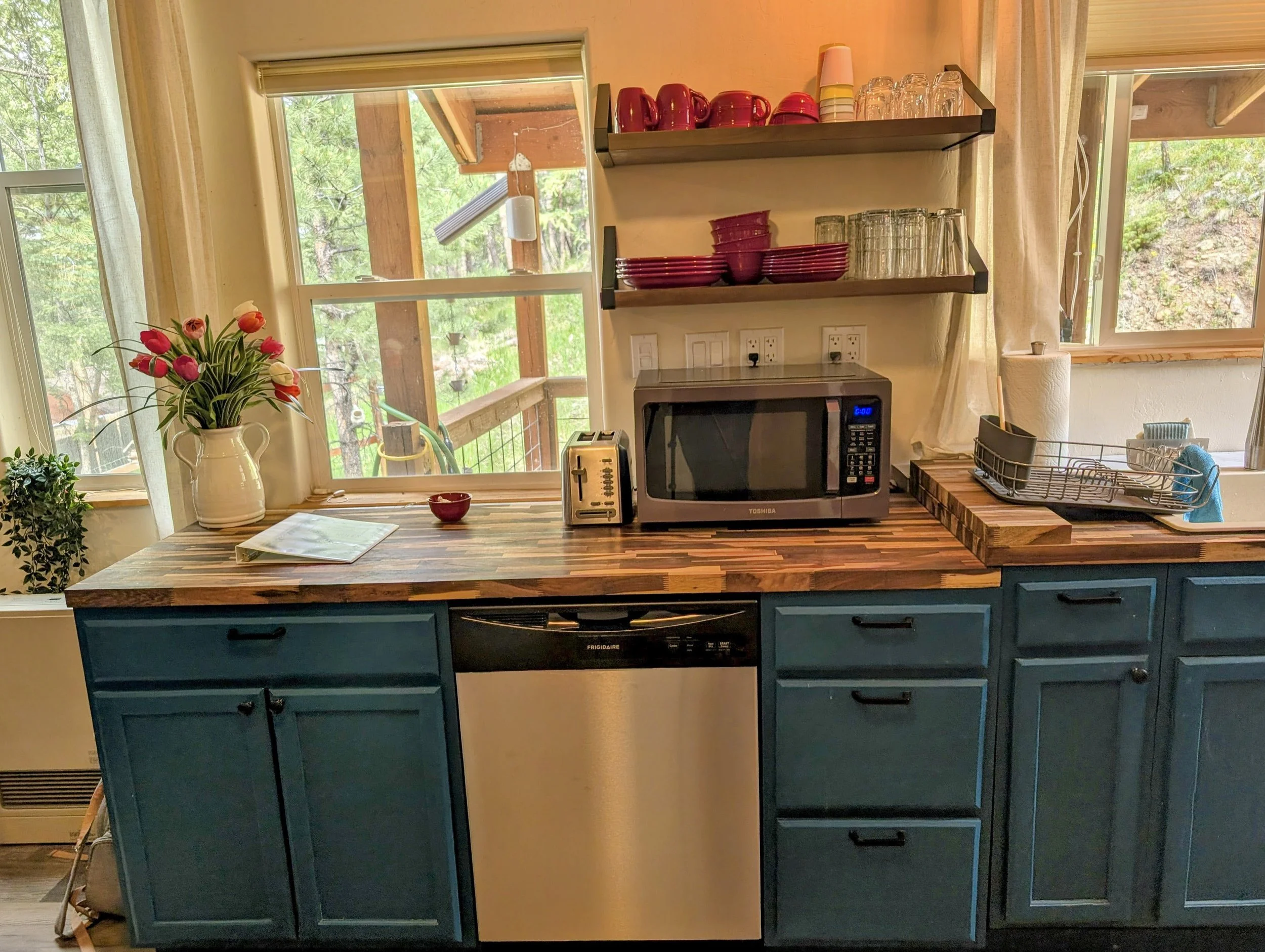 View of a kitchen counter with a vase of pink tulips, a microwave oven, a toaster, and dish drying rack near windows showing trees outside.