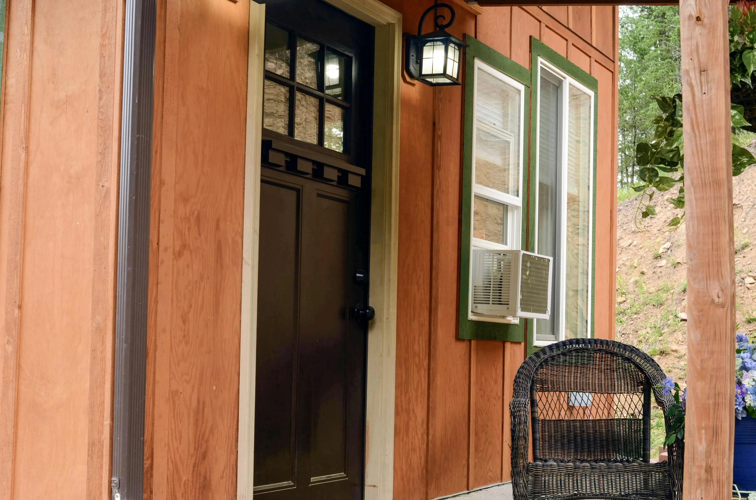 Front porch of a wooden house with a black door, green window shutters, and an outdoor light fixture. There is a wicker chair on the porch and ferns and flowers nearby.