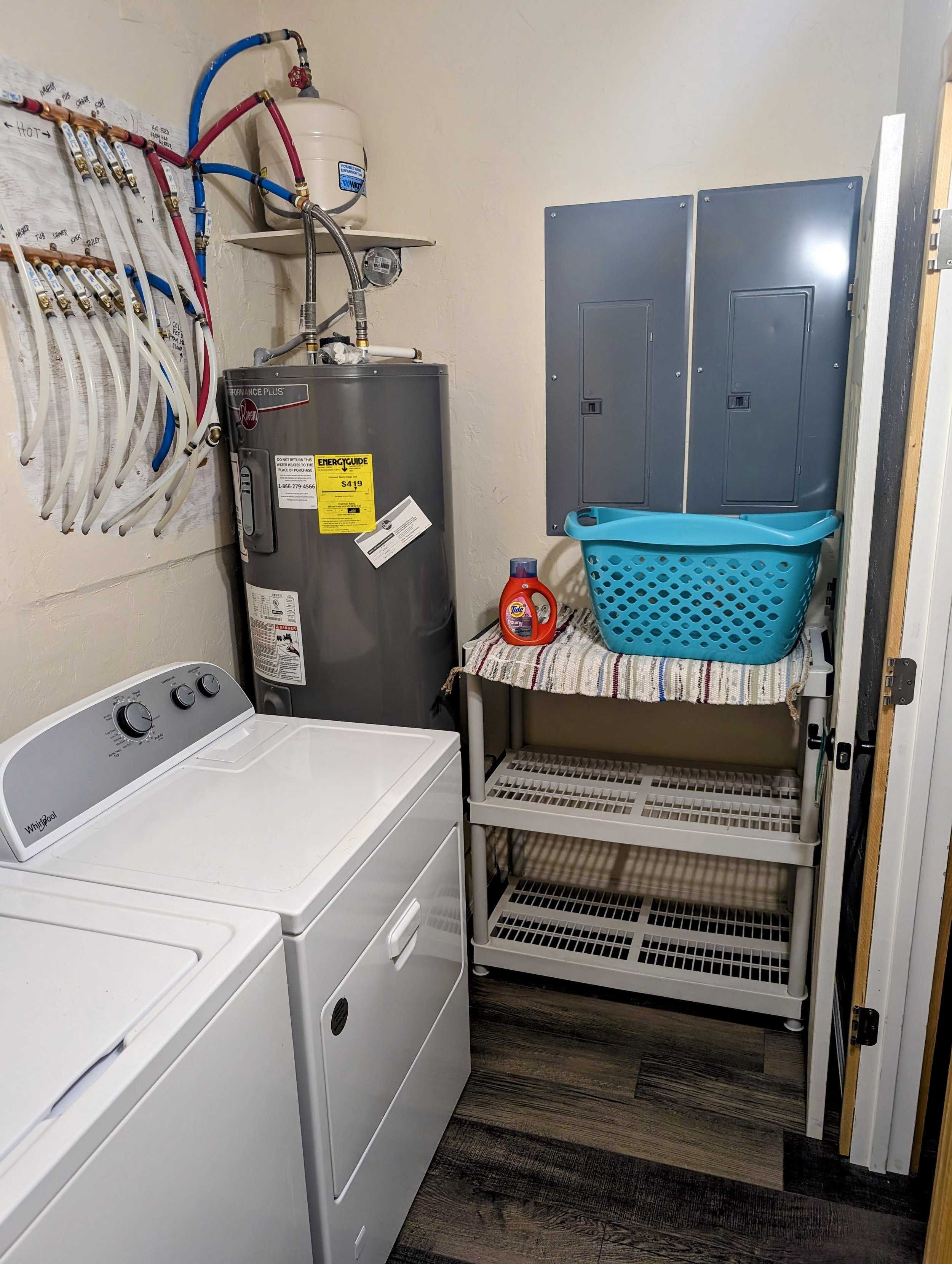 A laundry room with a white top-loading washing machine, a gray water heater, a blue laundry basket on a white shelving unit, a bottle of Tide detergent, and an electrical panel on the wall.