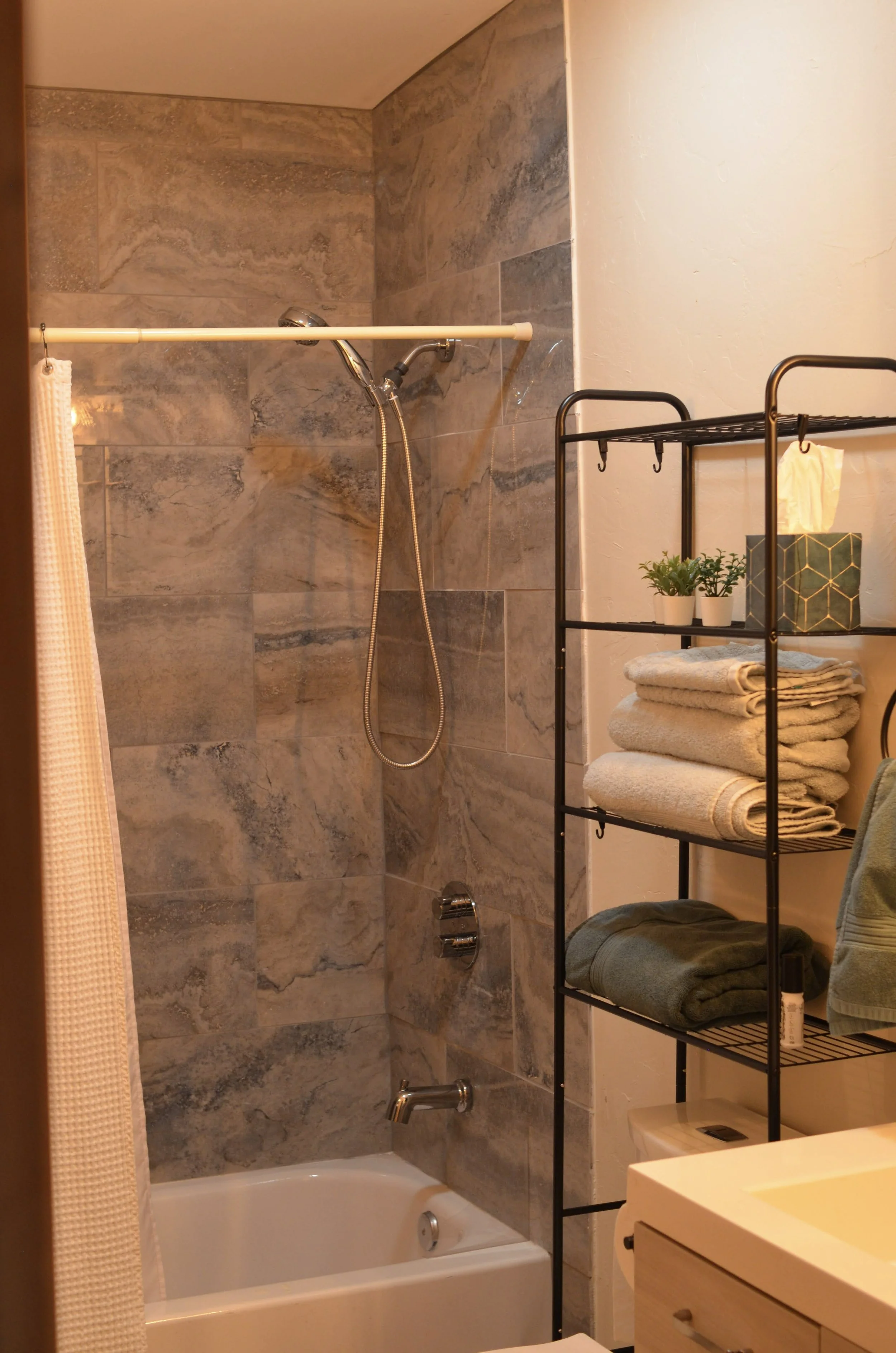 Bathroom with tiled shower area, a bathtub, a black metal shelving unit with towels and potted plants, a beige textured shower curtain, a sink with a wooden cabinet, and a toilet.