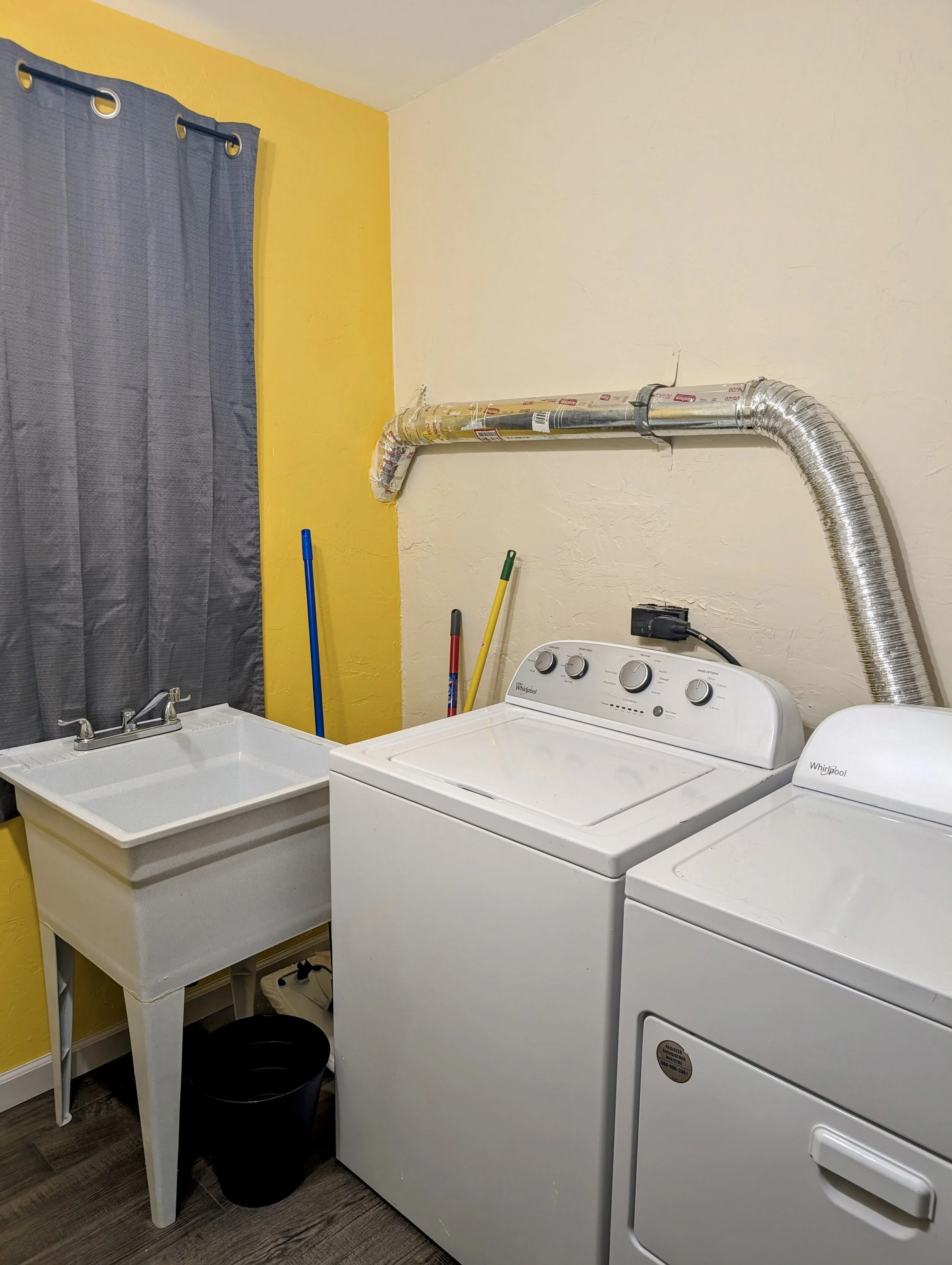 Laundry room with yellow and white walls, gray curtains, a utility sink, a washing machine, a dryer, and cleaning tools like mops and a bucket.