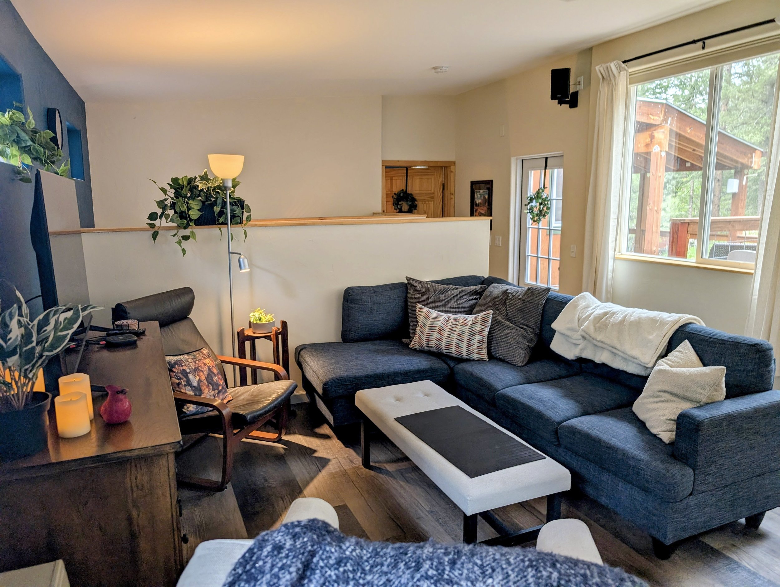 Living room with a sectional sofa, pillows, a wooden dining table with candles, houseplants, a window with curtains, and a view of a deck outside.