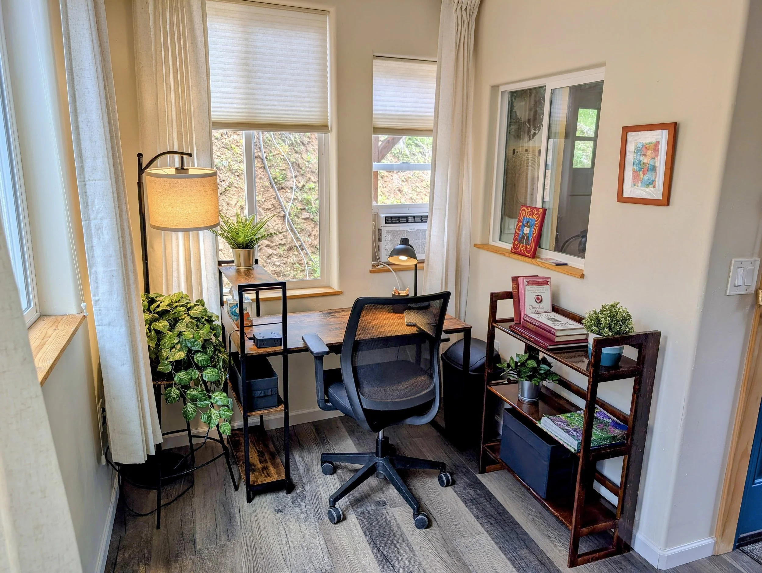 Home office corner with a wooden desk, black swivel chair, potted plants, books, artwork, and a window with blinds letting in natural light.