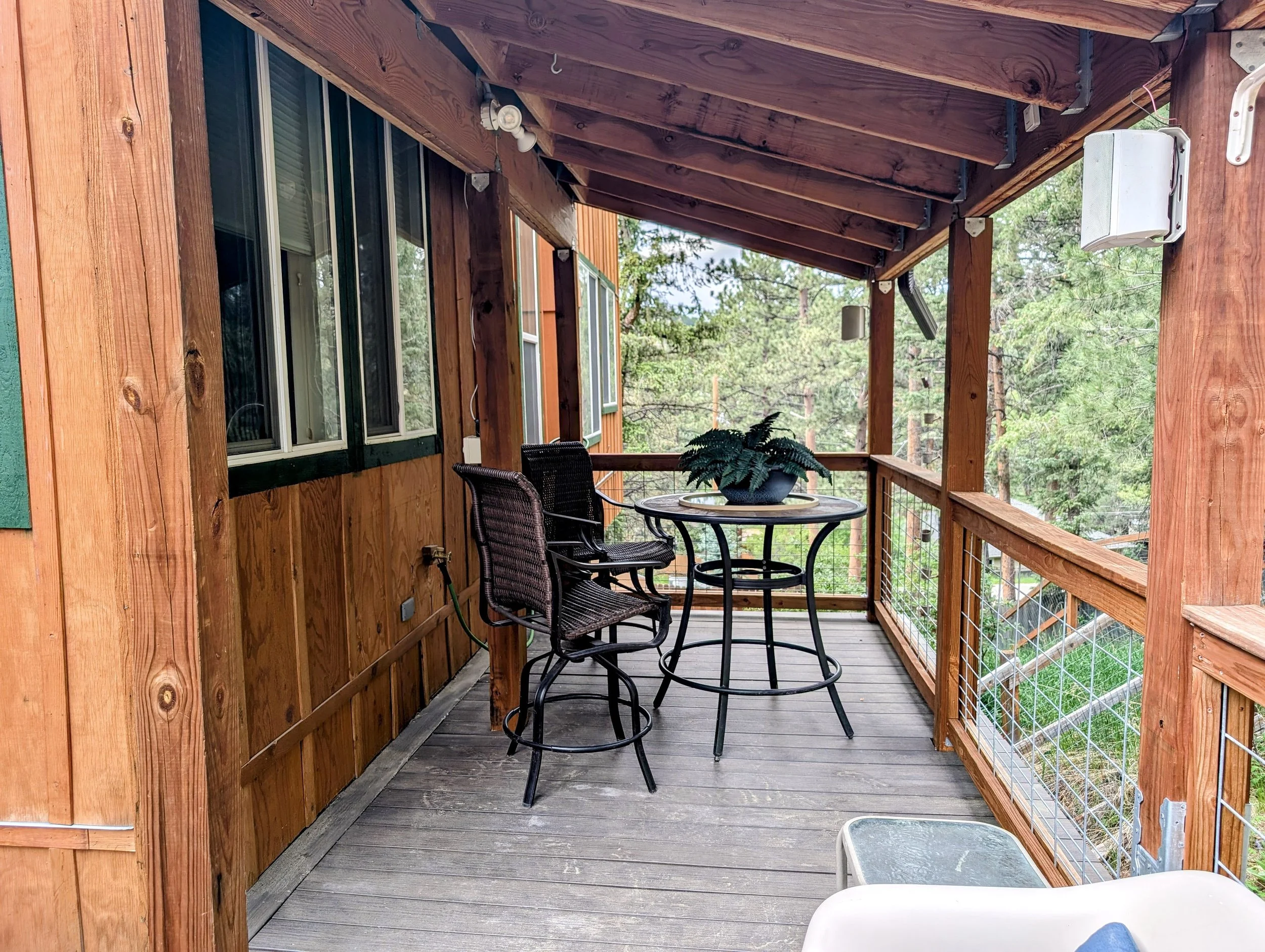 Wooden balcony with a black metal table, four black chairs, potted fern, surrounded by trees.