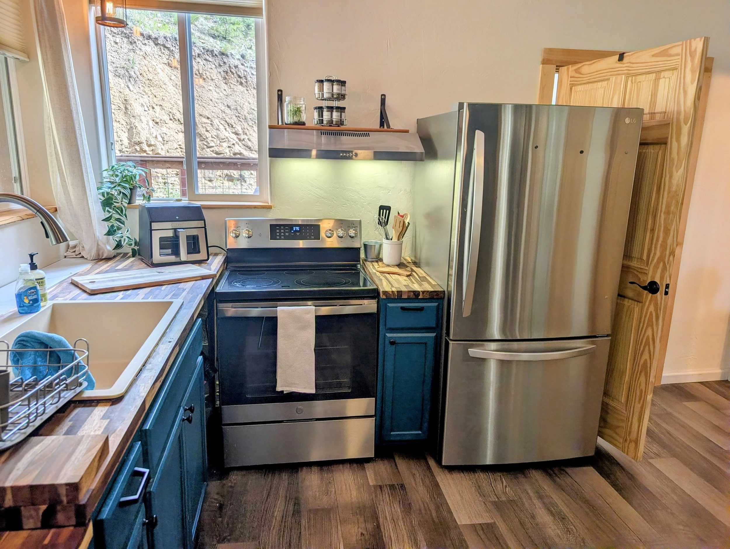 A cozy kitchen with wooden countertops, green cabinets, stainless steel appliances, a window with a view of a rocky hillside, a potted plant, and various kitchen utensils and appliances.