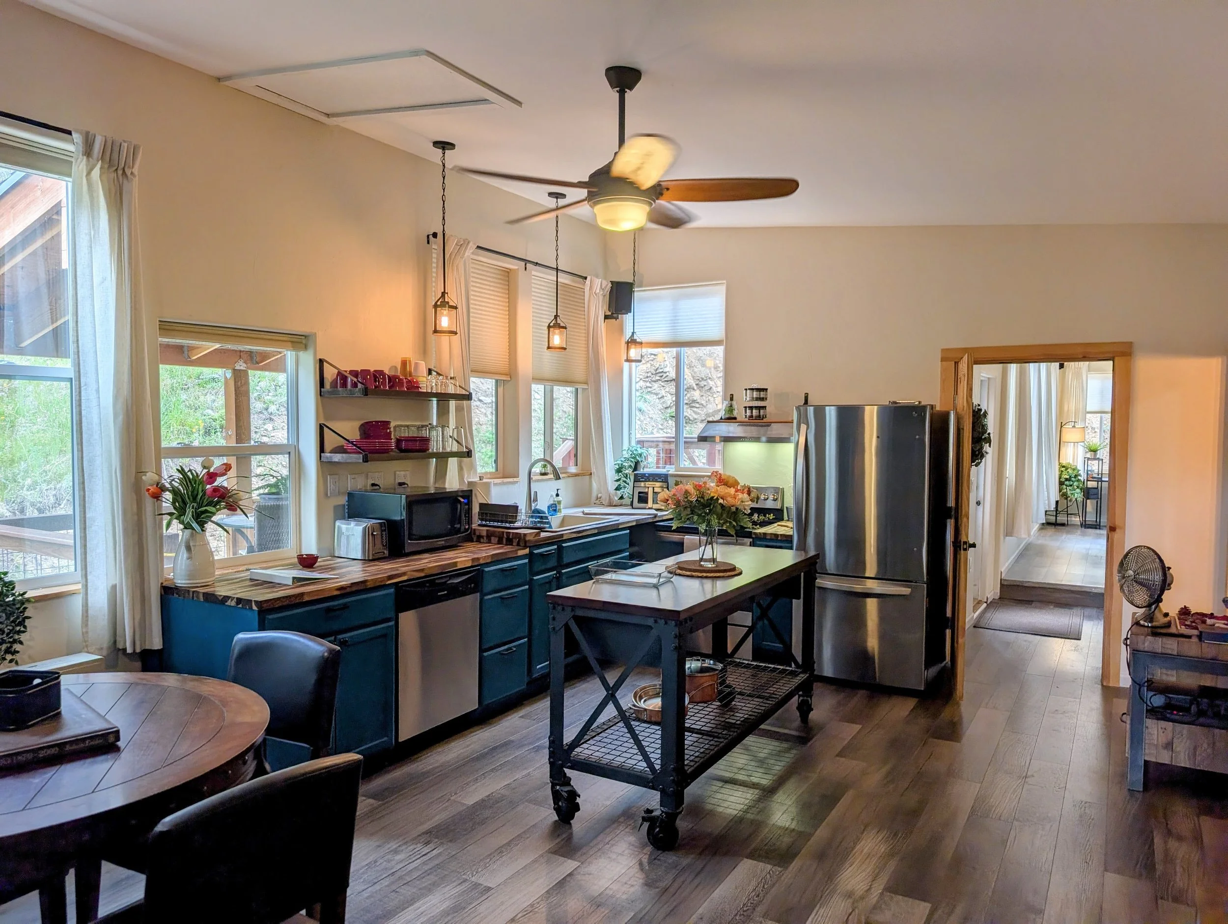 Interior view of a kitchen with large windows, pendant and ceiling lights, a ceiling fan, plants, flowers, a refrigerator, and kitchen appliances.