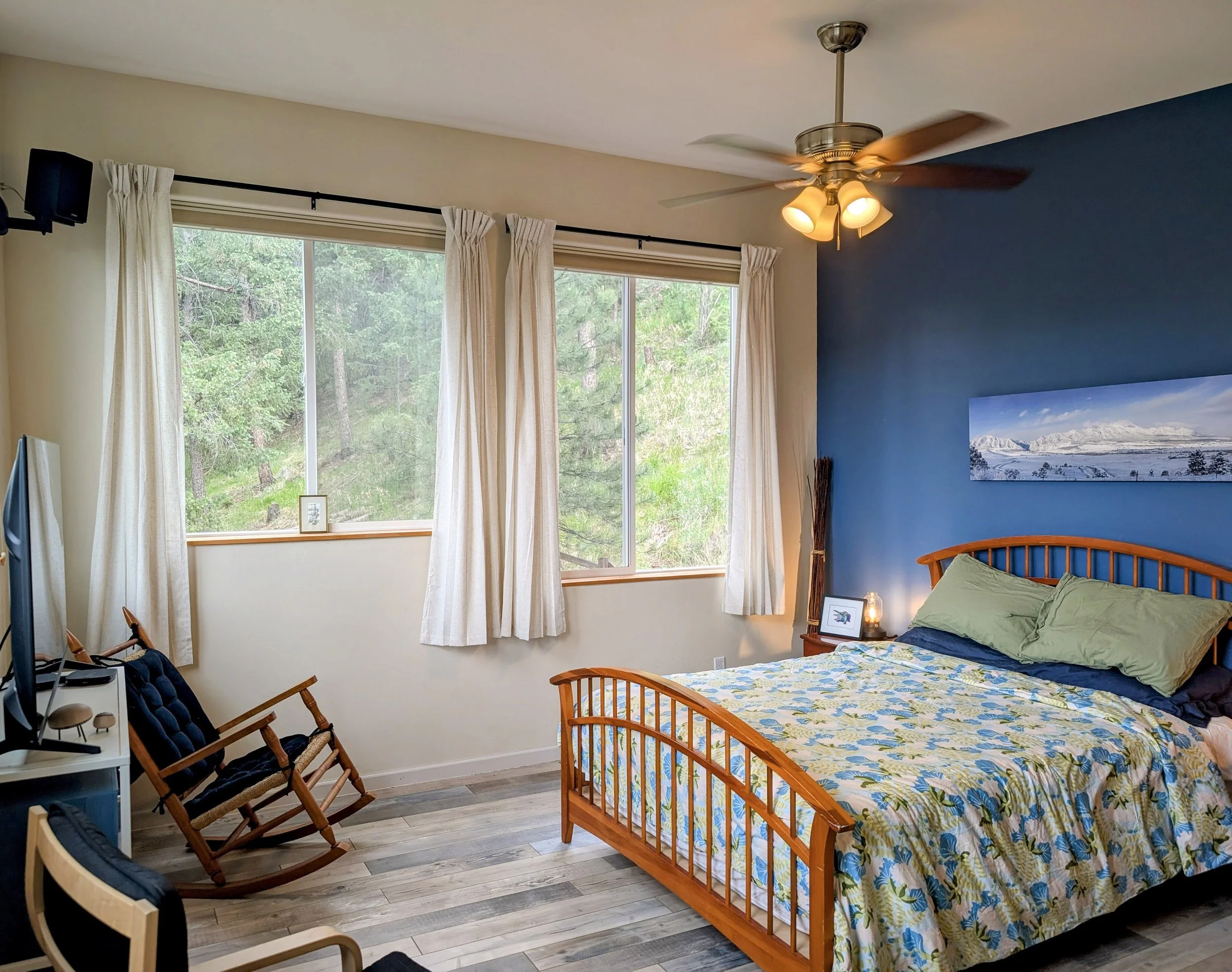 A bedroom with large windows showing trees outside, a wood bed with a colorful floral quilt and green pillows, a blue accent wall with a landscape painting, ceiling fan with lights, and various chairs and furniture.