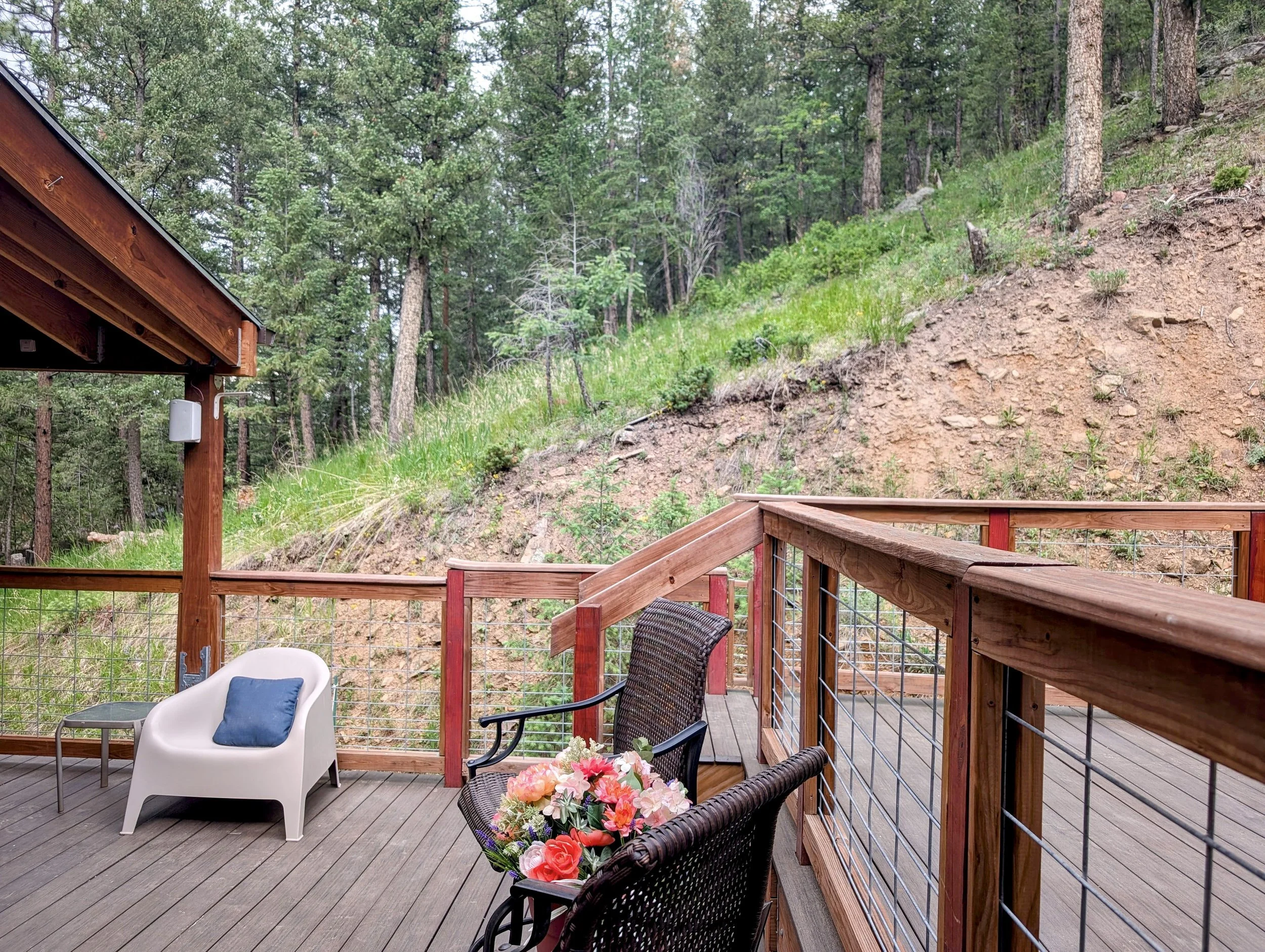 A wooden deck with outdoor furniture including a white chair with a blue pillow, a wicker chair with pink flowers, and two other chairs, overlooking a forested hillside with tall pine trees.