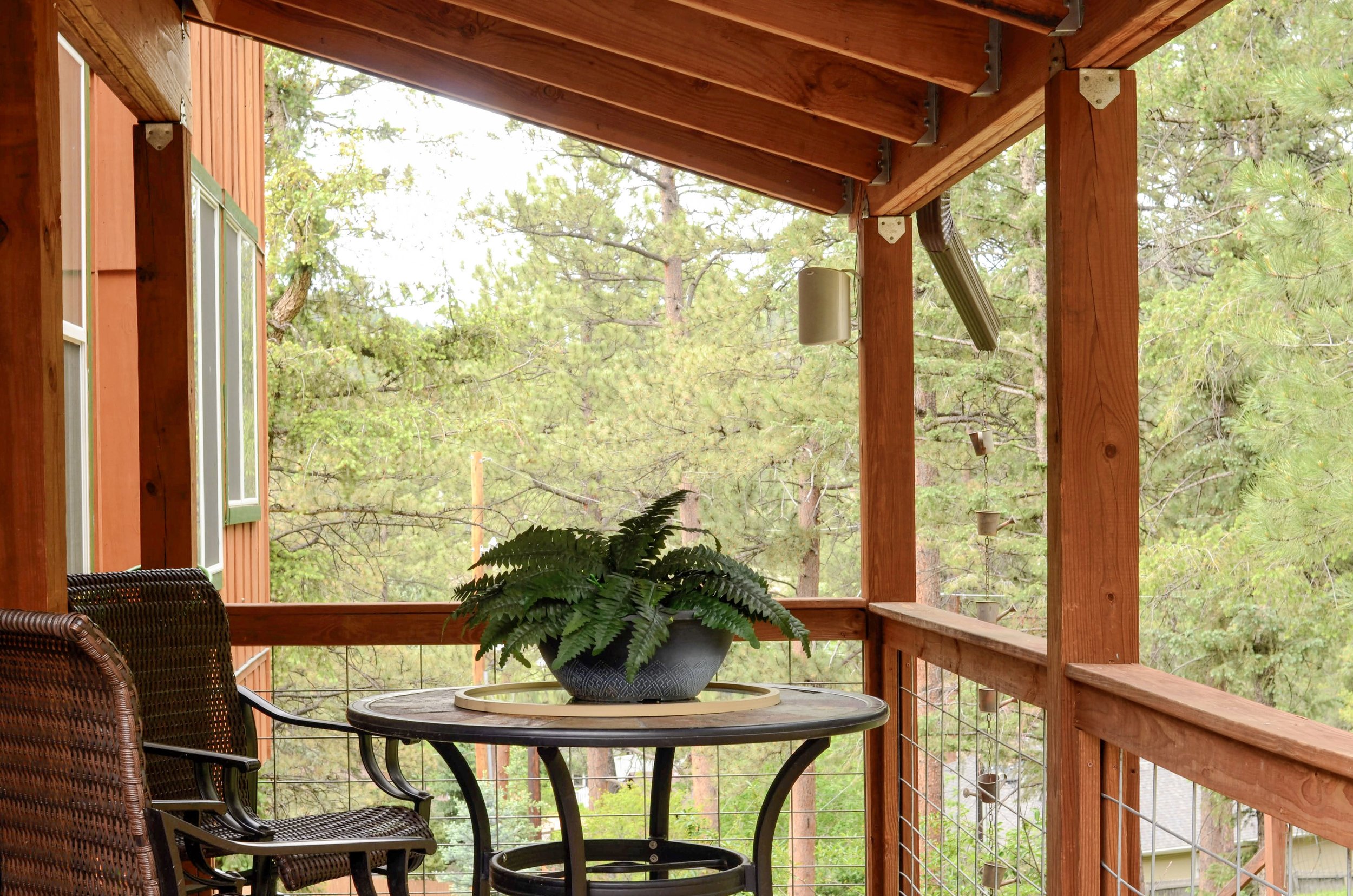 A wooden deck with a round table and two chairs, and a potted fern plant on the table. The deck is surrounded by a railing and is attached to a house with orange walls and green window trim. In the background, tall pine trees看到