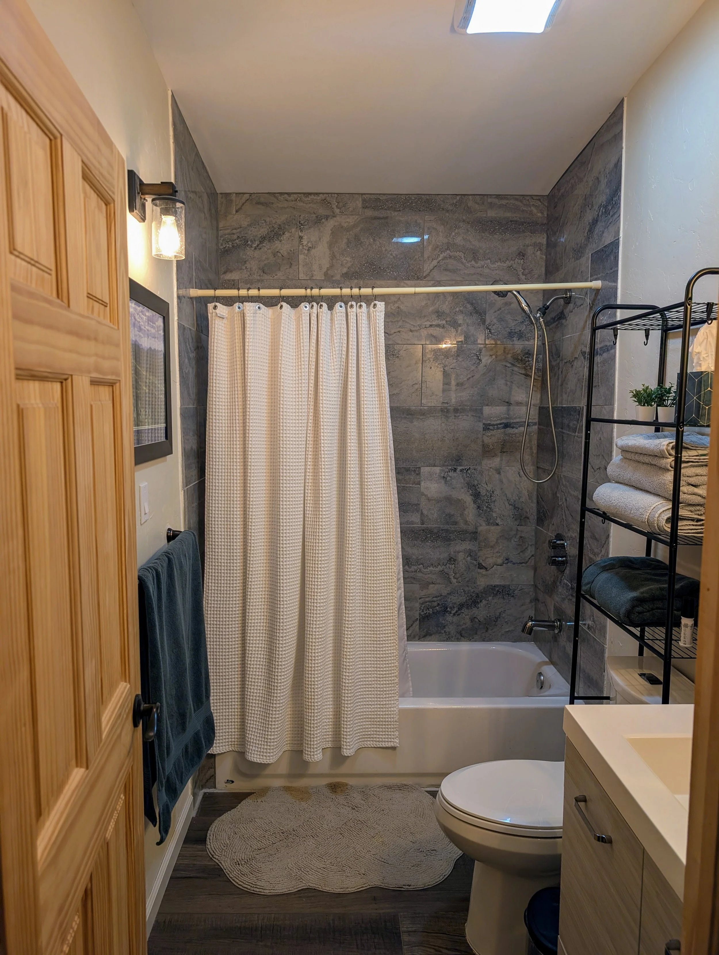 Bathroom with a bathtub and shower, beige and gray tiles, a toilet, a wooden cabinet, a black towel rack with blue towels, a small black shelf with folded towels and a potted plant, a beige rug, and a ceiling light.