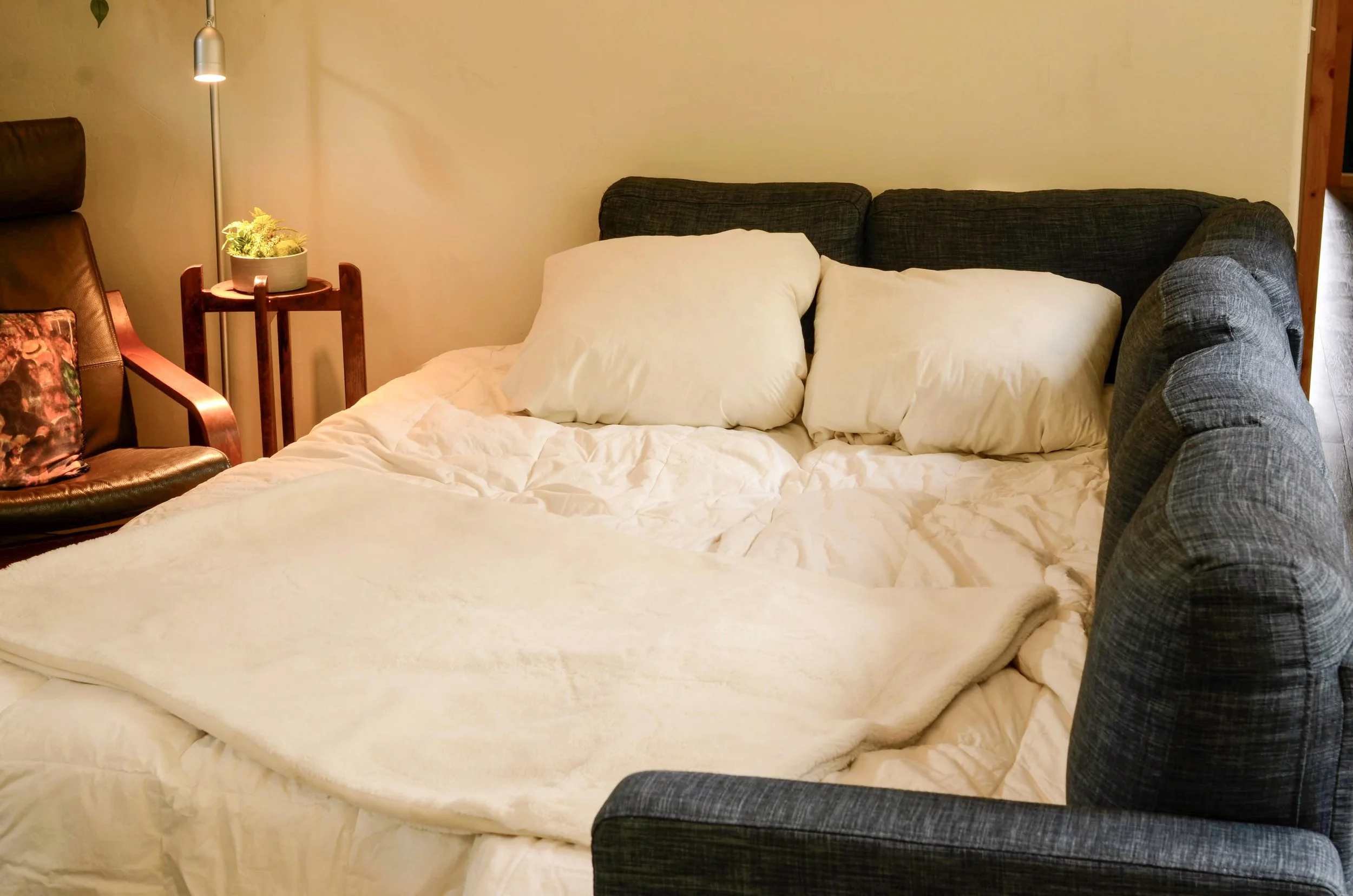 A cozy bedroom scene with a bed, pillows, a wooden side table with a plant, a floor lamp, and a dark-colored armchair with a floral pillow.