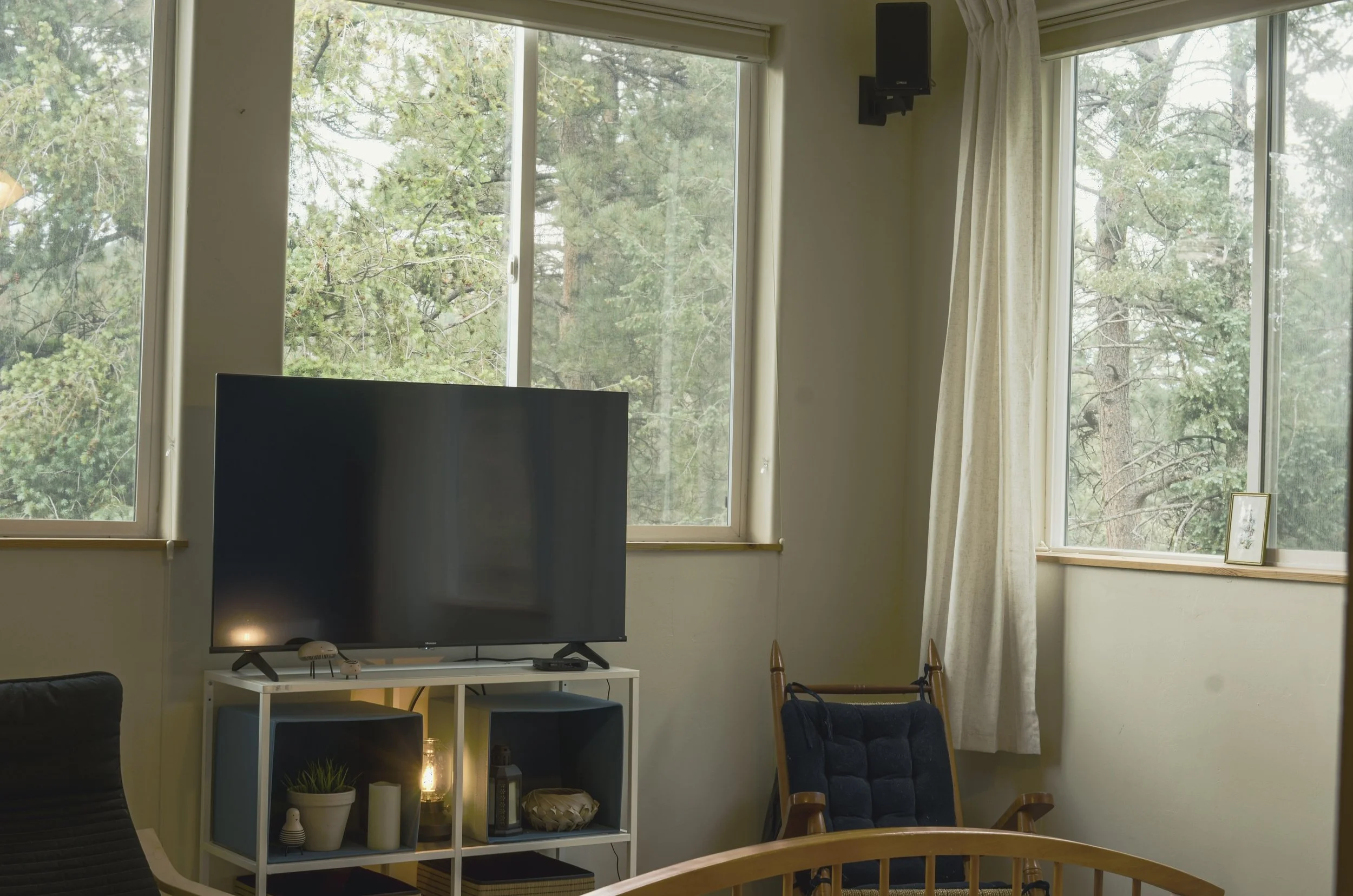 Living room with large windows showing trees outside, a flat-screen TV on a white stand, a dark sofa, a patterned armchair, and curtains on either side of the windows.