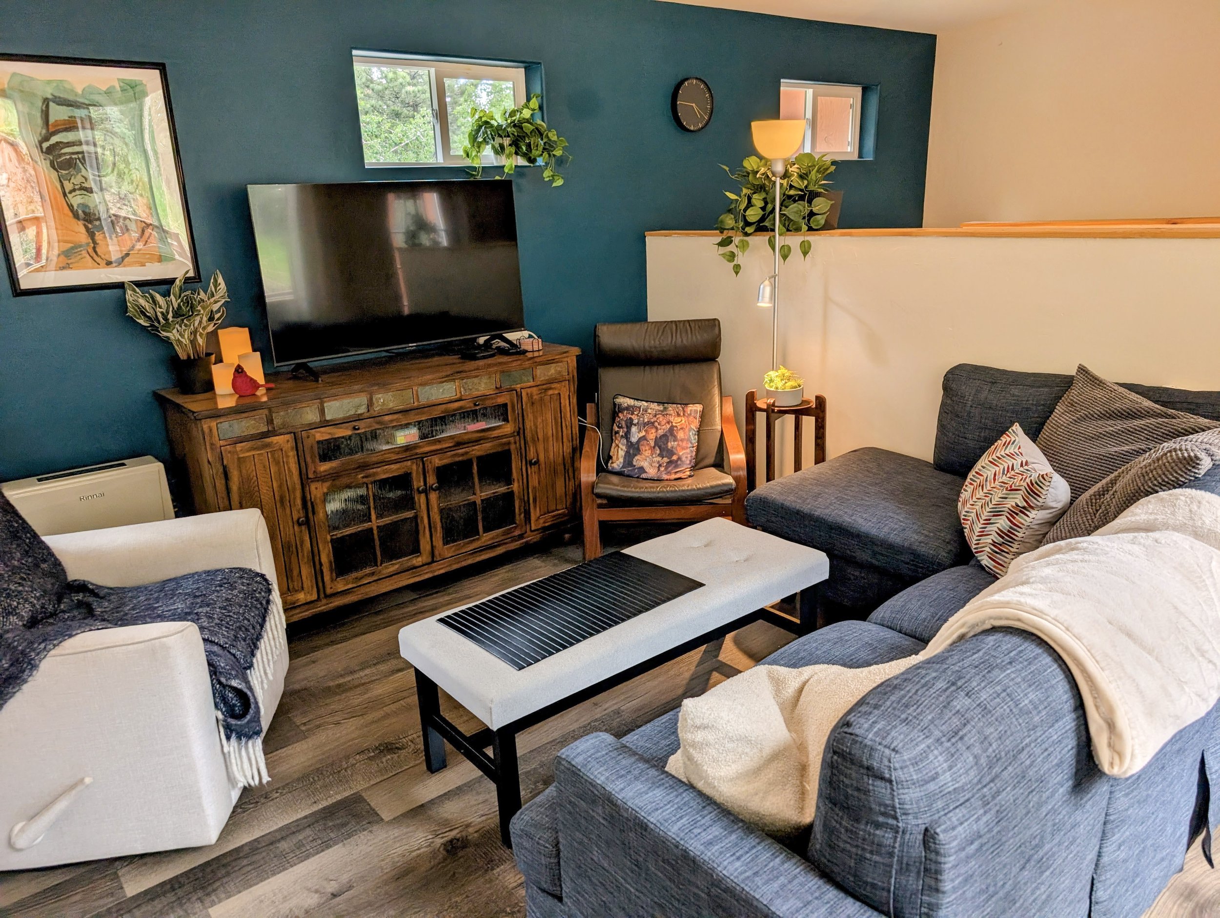 A cozy living room with blue accent wall, a large flat-screen TV on a wooden cabinet, a black leather armchair with a decorative pillow, a white sofa with beige and patterned pillows, a coffee table, a tall floor lamp with a globe-shaped shade and a 