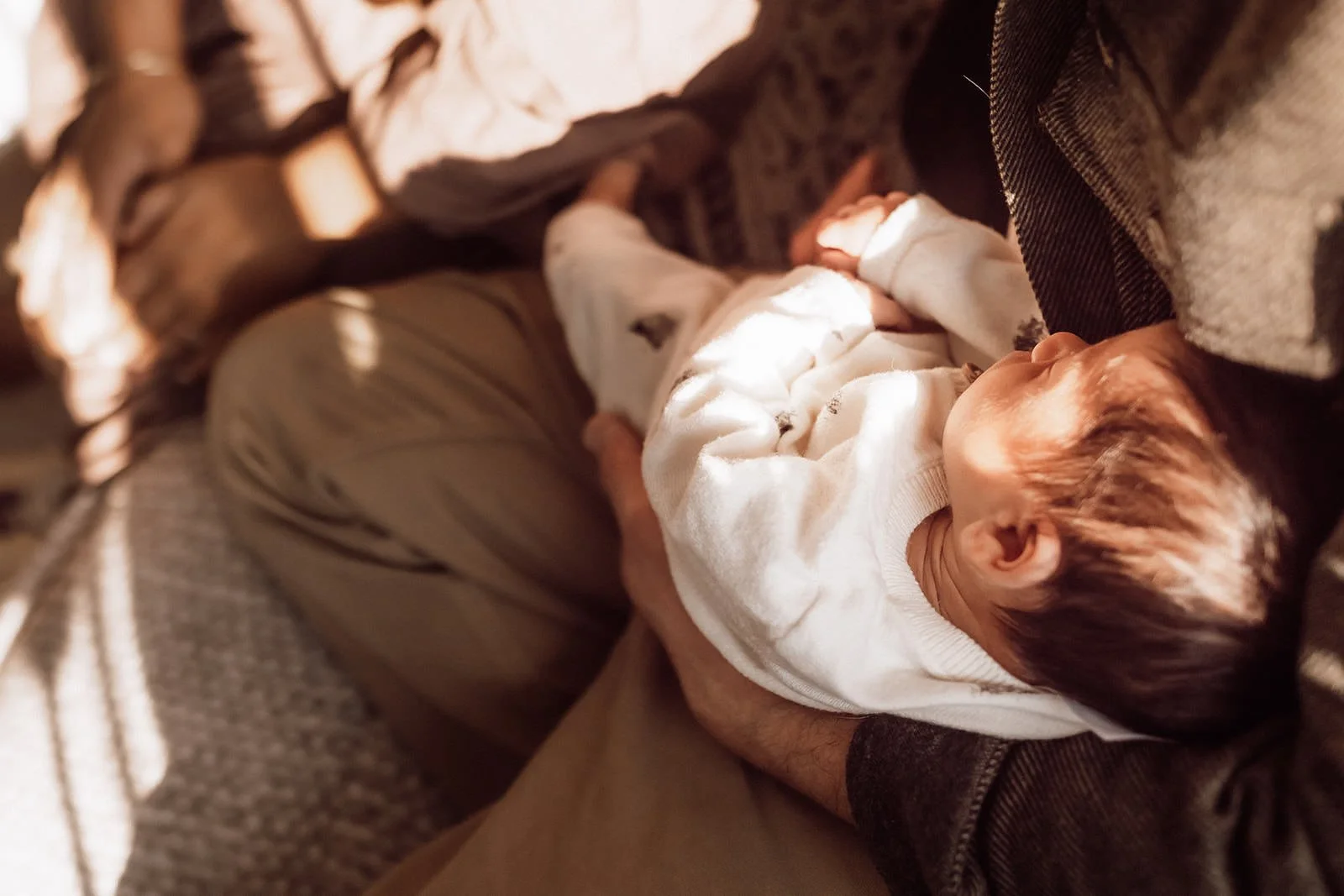 A young child wearing a white sweater lying on an adult's lap with a hand gently resting on their stomach, outdoors in warm sunlight.