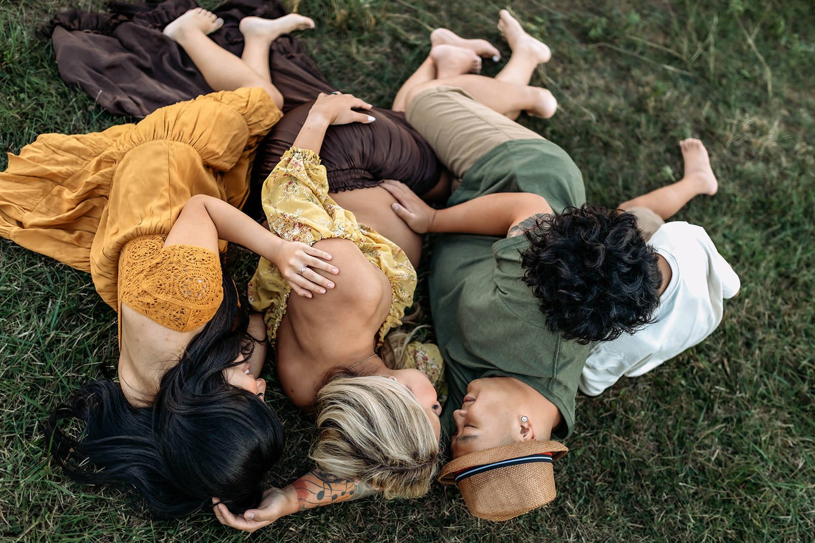 Four people lying on the grass embracing each other, two women and two men, wearing casual summer clothes, one woman with black hair, one woman with blonde hair, one man with dark curly hair, and one man wearing a tan fedora.