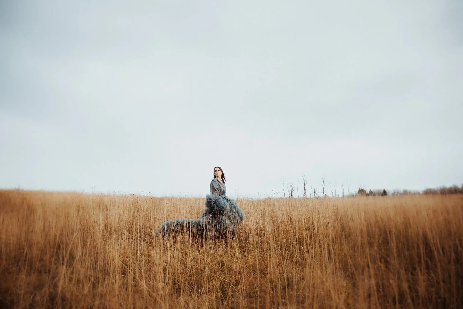 A woman in a gray dress sits on a large gray rock in a field of tall, dry grass under a cloudy sky, with sparse trees in the distance.