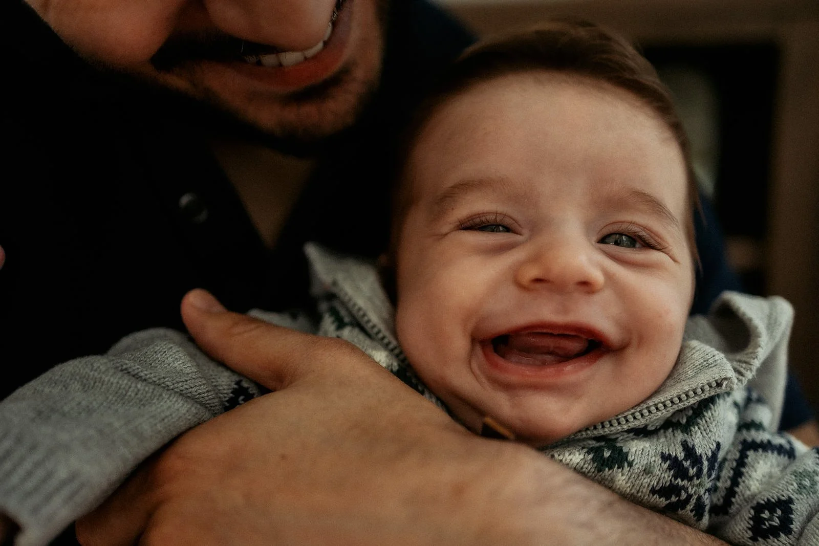 A person holding a smiling baby with blue eyes and light brown hair, wearing a gray and white patterned sweater.