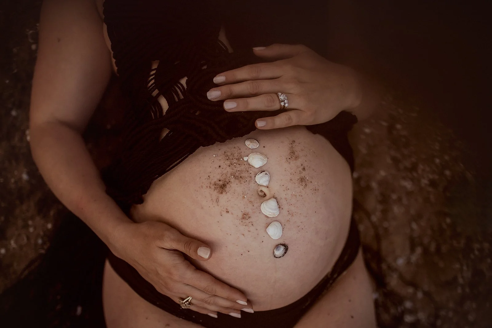 Close-up of a pregnant woman's belly with seashells arranged along the center, sand sprinkled on the skin, and hands resting on the belly, one hand wearing a silver ring with multiple stones.