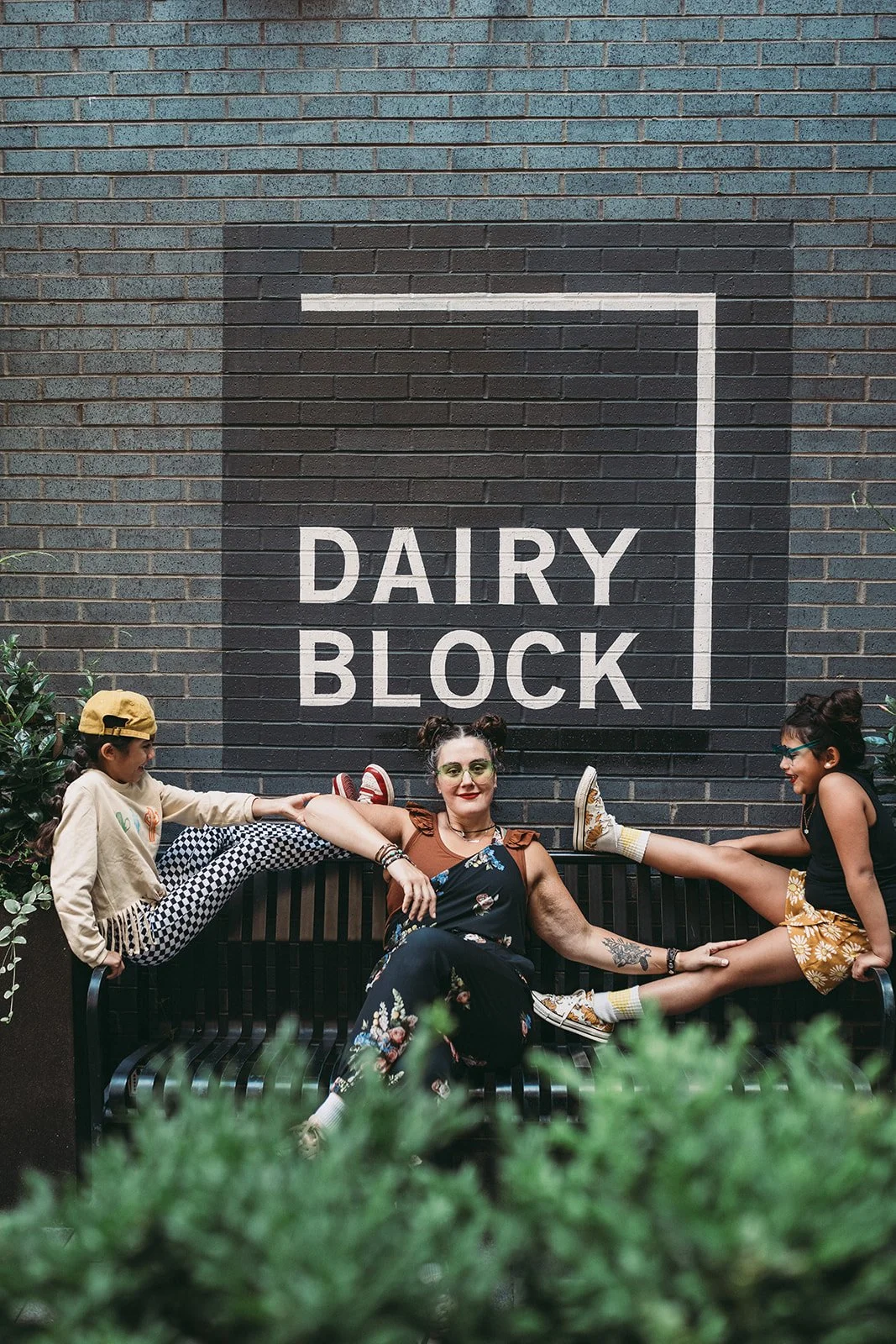 Three people sitting on a black bench in front of a wall with a large sign that says "Dairy Block." Two children are playfully stretching their legs and holding hands, one girl wearing a yellow cap, beige hoodie, and checkered pants, and another girl wearing sunglasses, a black tank top, and yellow floral shorts. An adult woman is sitting in the middle, smiling, with her legs stretched out and her hand holding the child's leg.