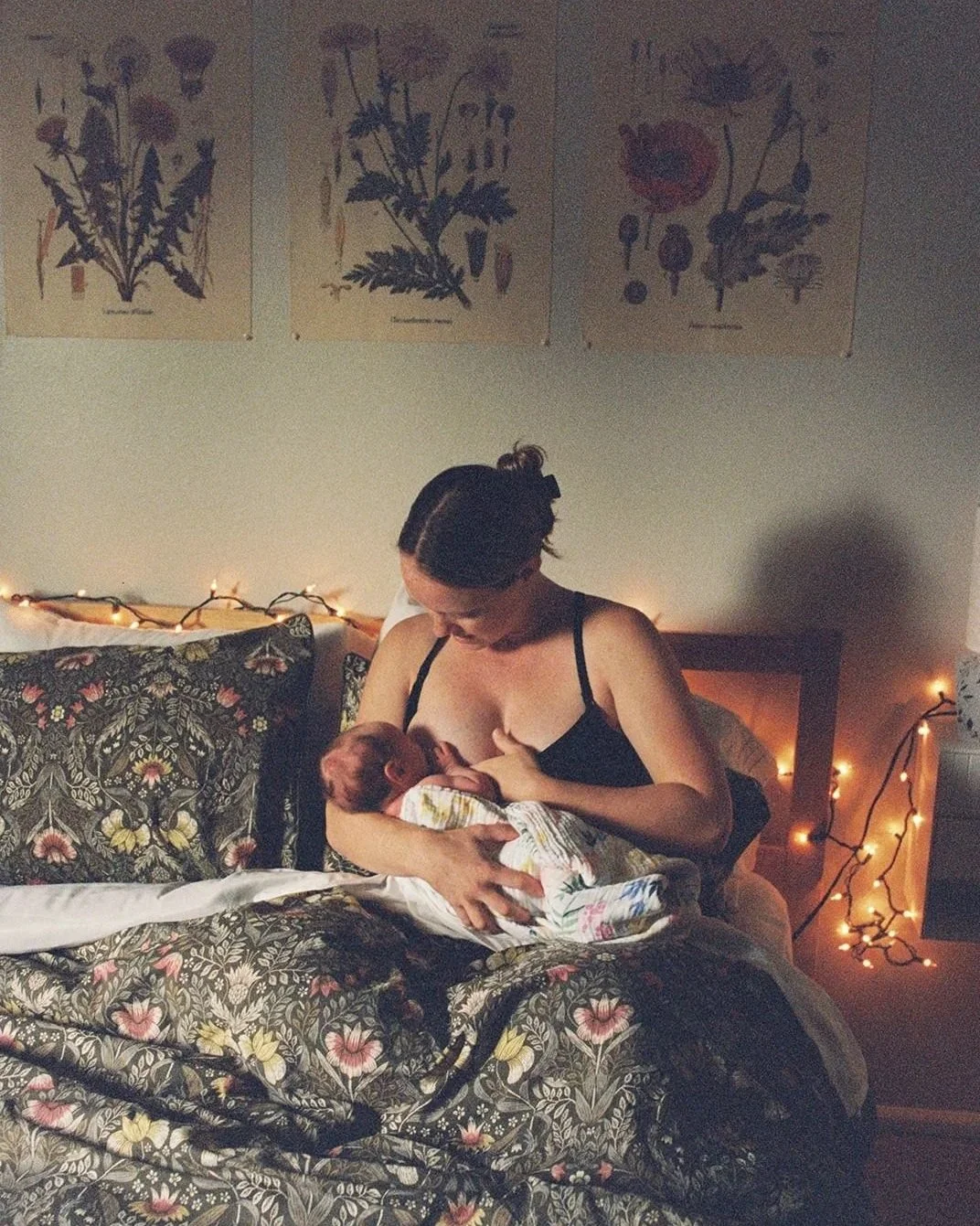 A woman breastfeeding a newborn baby on a bed decorated with floral bedding and string lights, with botanical prints on the wall.
