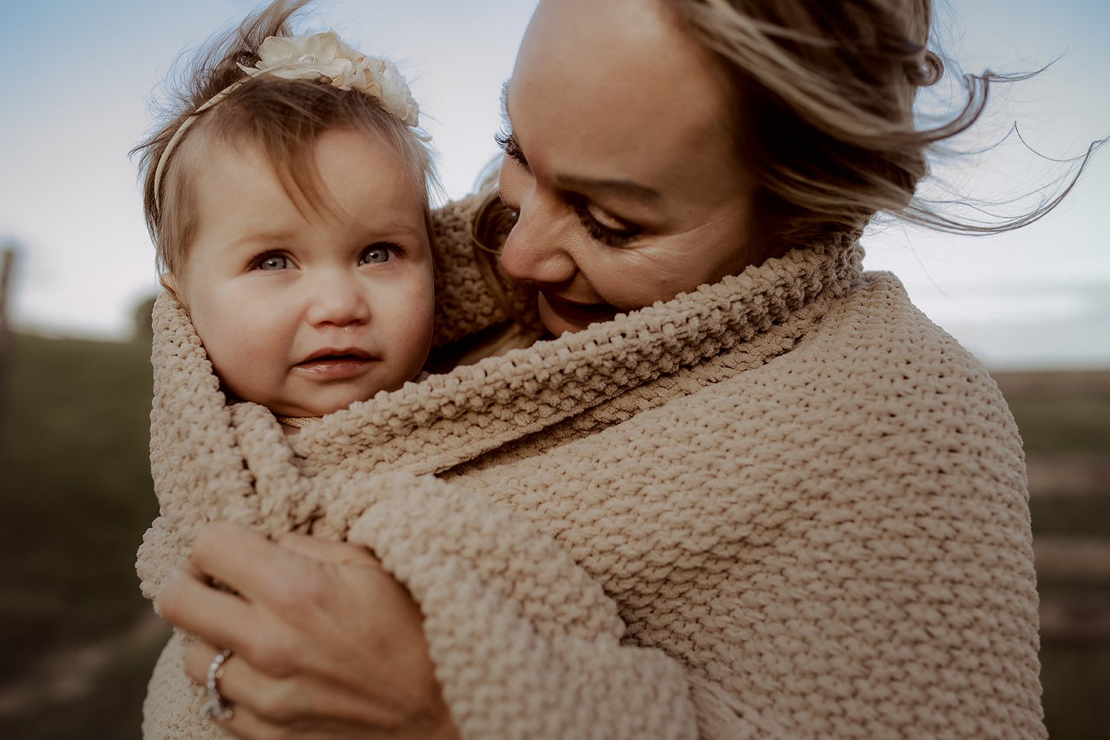 A woman wrapped in a beige knitted blanket holding a young girl with a cream headband in an outdoor setting with a cloudy sky.
