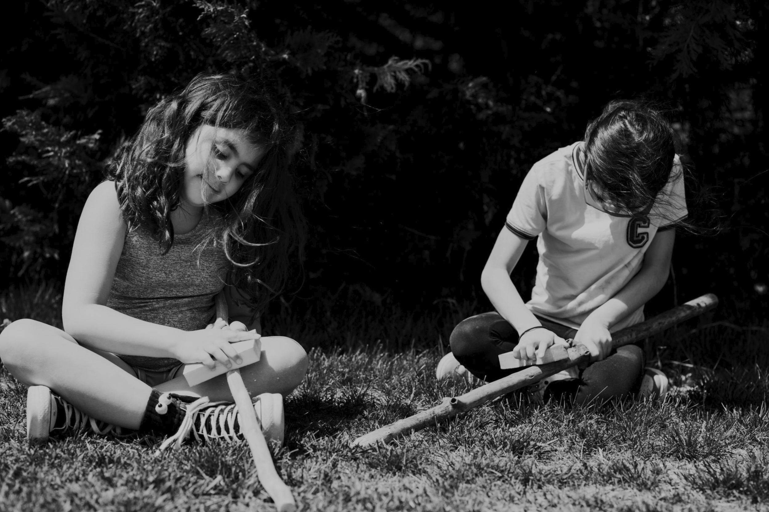 Two girls sitting on grass outdoors in black and white, using tools and paper, with trees in the background.
