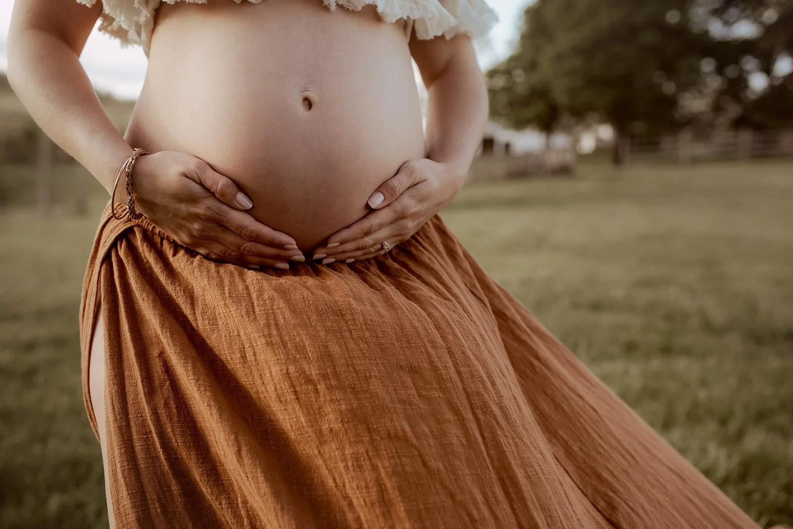 Close-up of a pregnant woman cradling her belly outdoors, wearing a brown skirt and a white top, with trees and a fence in the background.