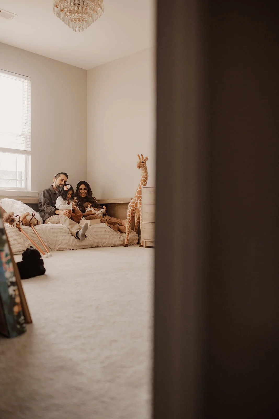 A family sitting on a bed in a bright, cozy room with a large window, a giraffe plush toy, and some stuffed animals, as seen from behind a partially closed door.