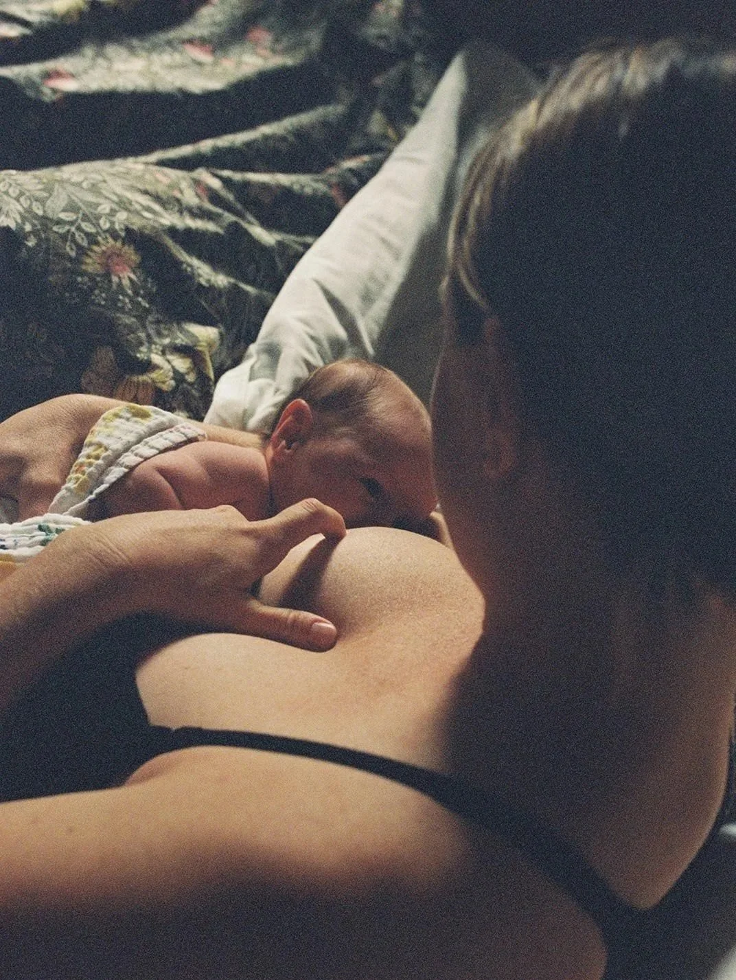 A woman lying on a bed with a pillow, gently holding and kissing a newborn baby who is resting on her chest, with a dark floral bedspread in the background.