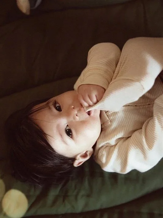A young child with dark hair lying on a dark-colored surface, looking up at the camera, with one finger in their mouth.
