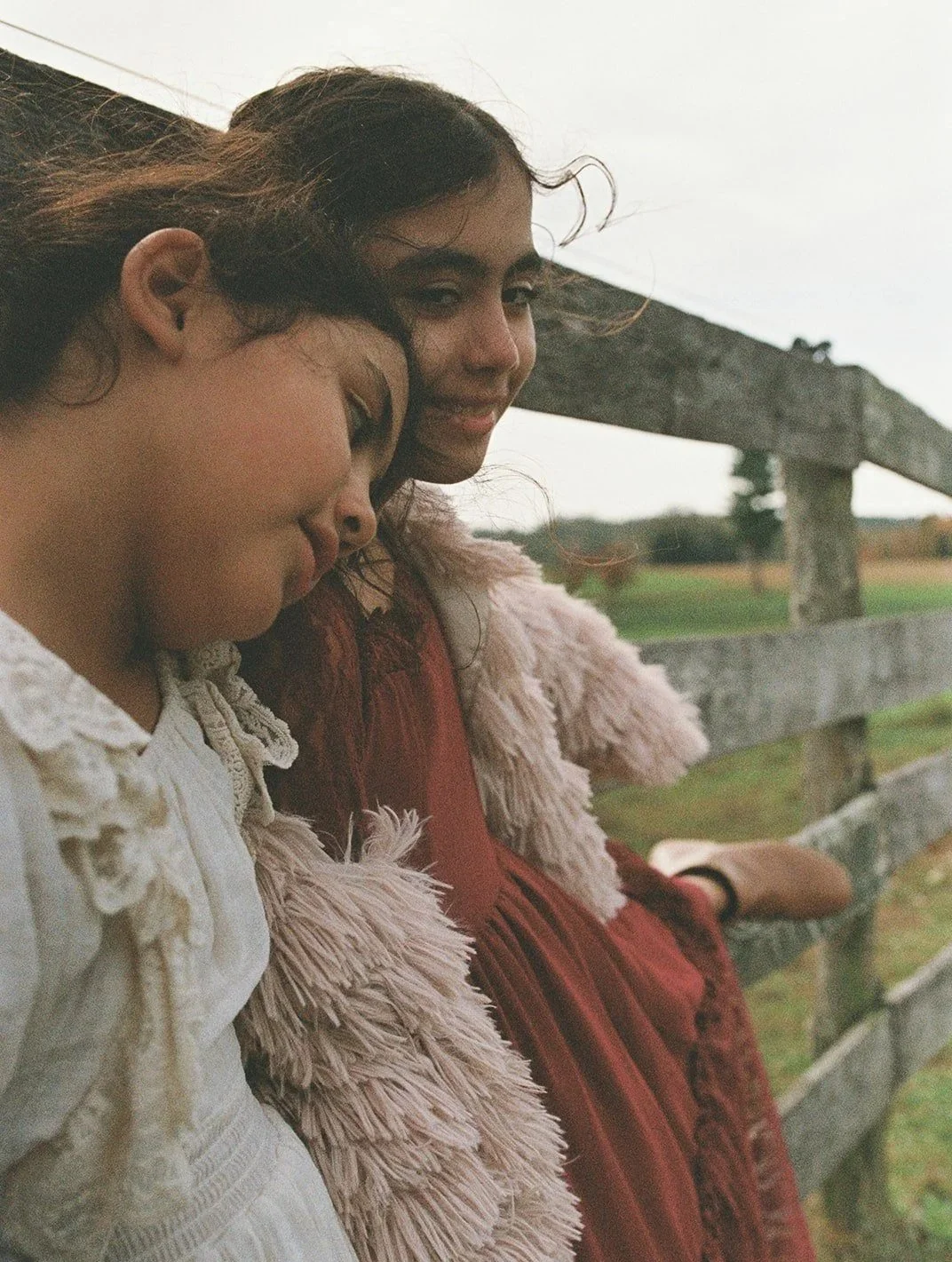 Two women sitting on a wooden fence outdoors during dusk, close together with one resting her head on the other's shoulder, both with relaxed and content expressions.