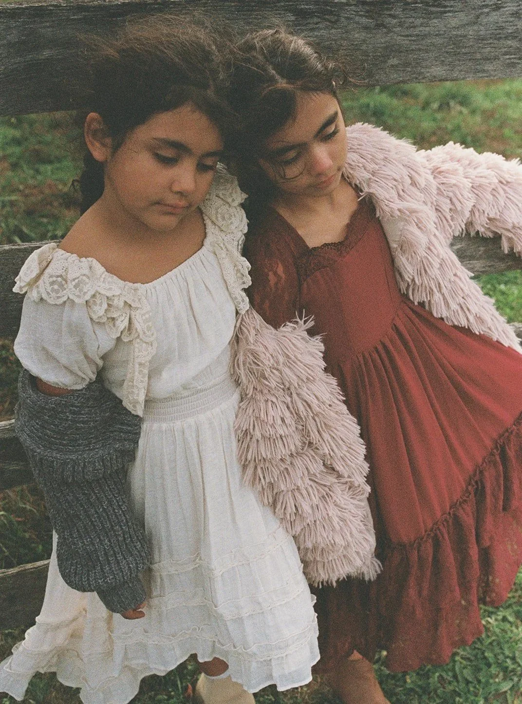 Two girls sitting on a wooden bench outdoors, leaning against each other with closed eyes.