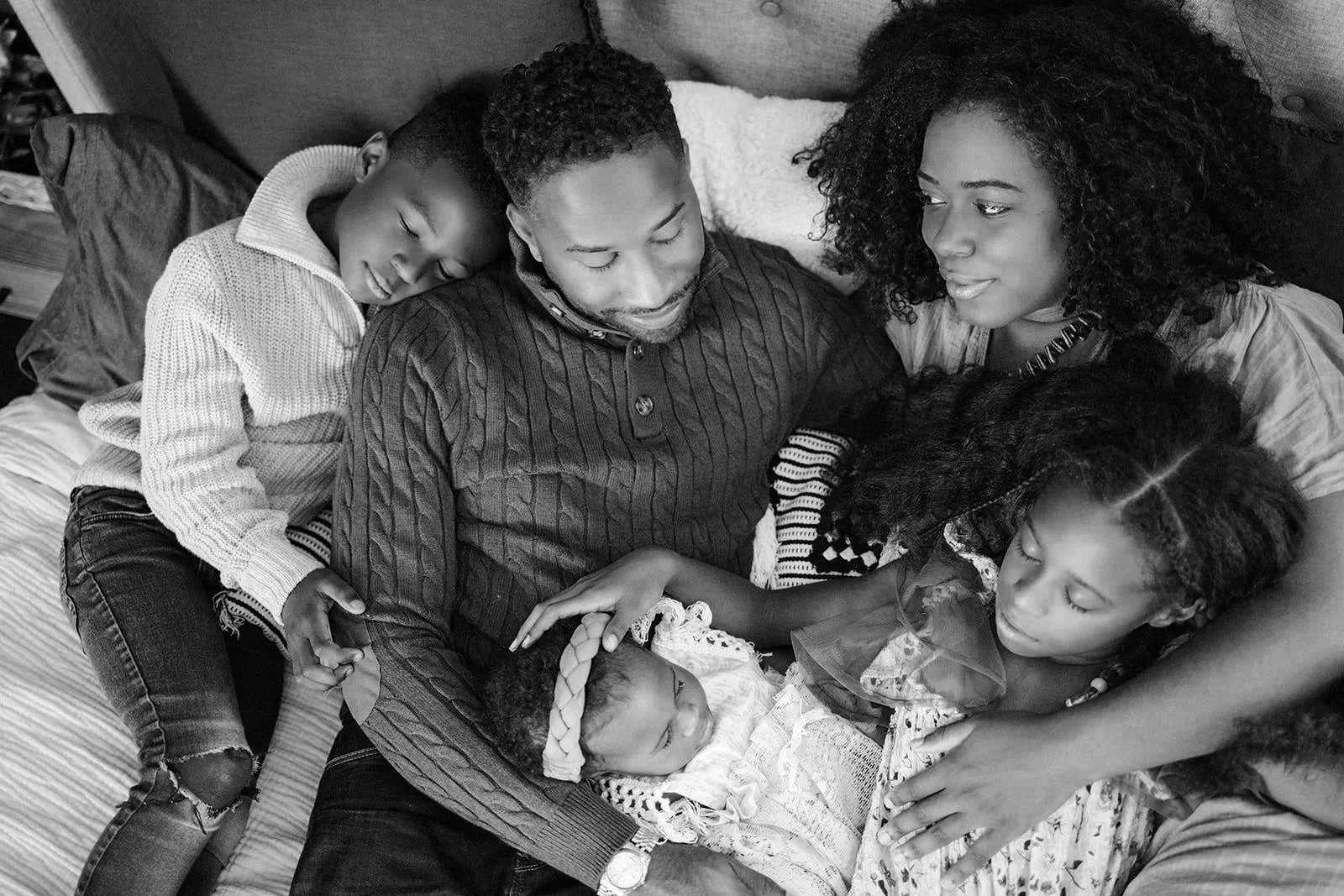 A family of five African Americans gathered on a bed, looking at a newborn baby girl, celebrating a new addition to their family.