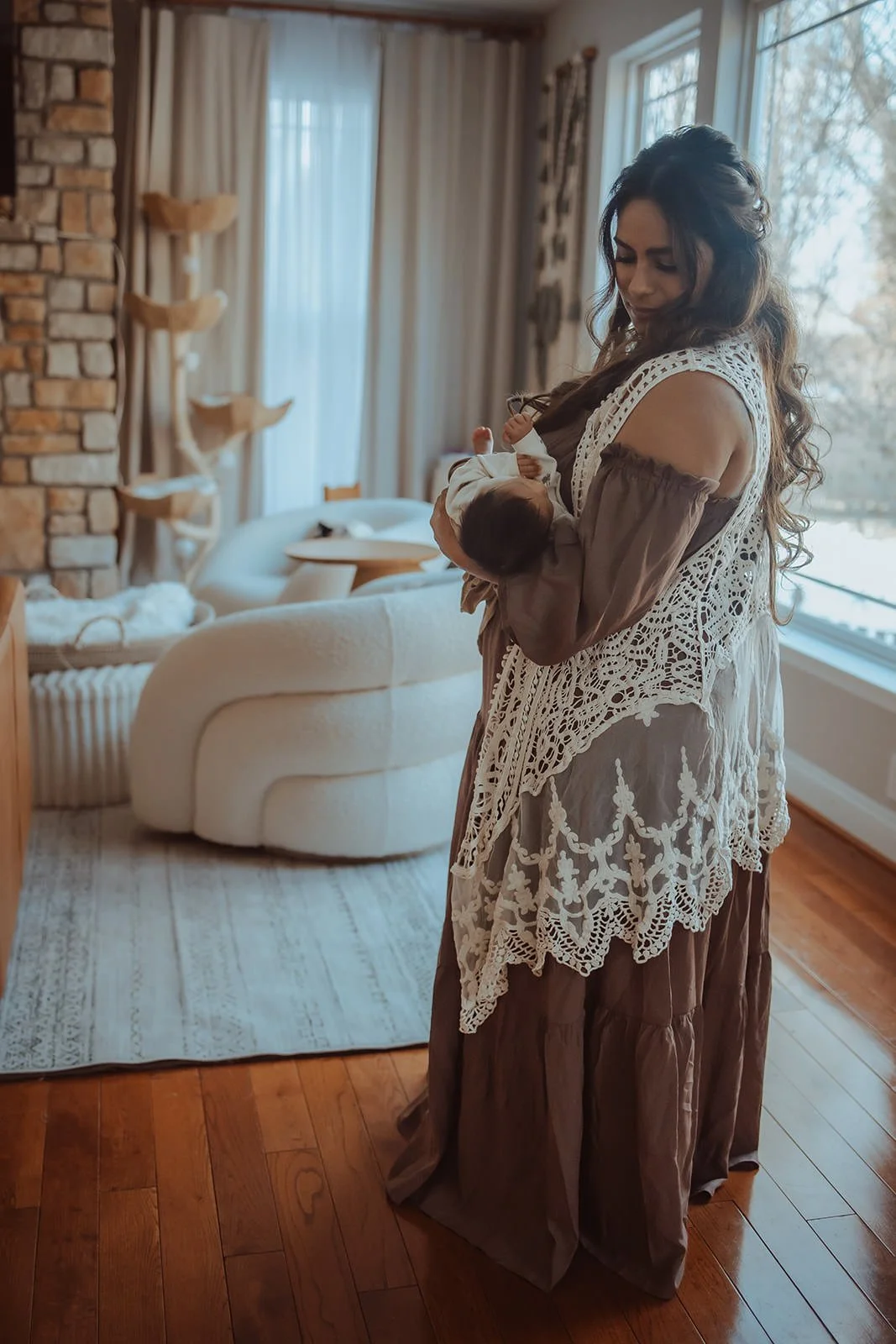 A woman holding a newborn baby indoors near a window, with a cozy living room in the background.
