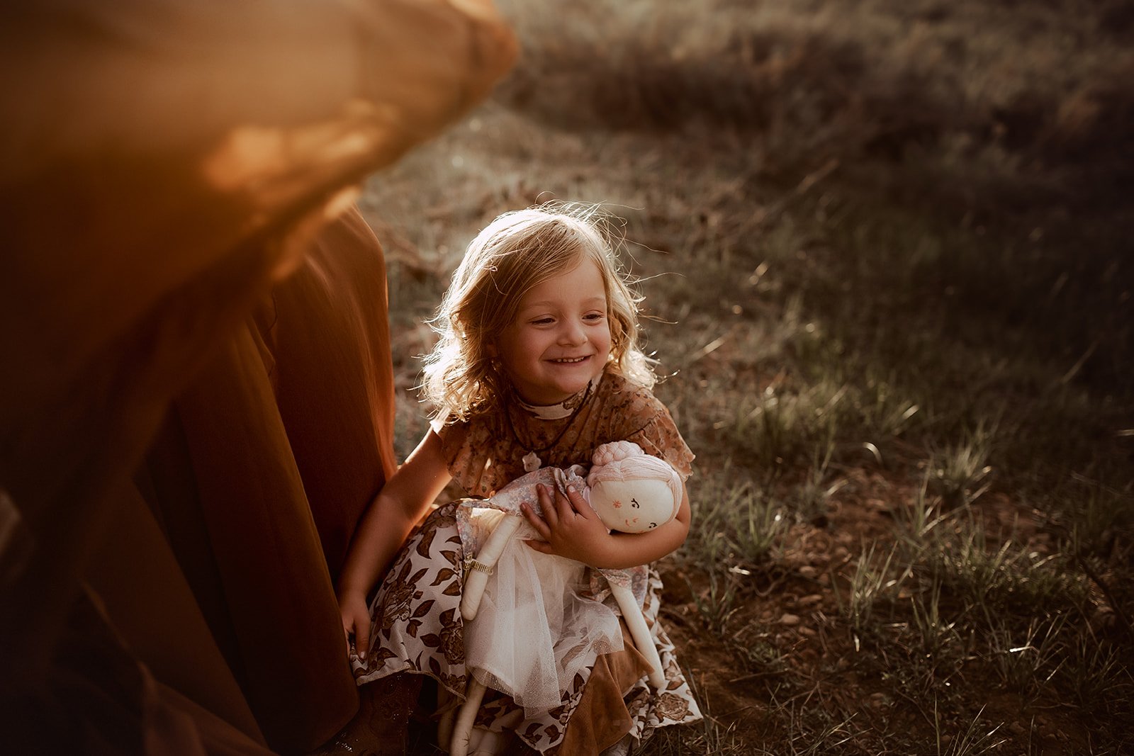 A young girl with blonde hair smiling while sitting outdoors on the ground, holding a doll. The sunlight illuminates her hair, and she appears happy.