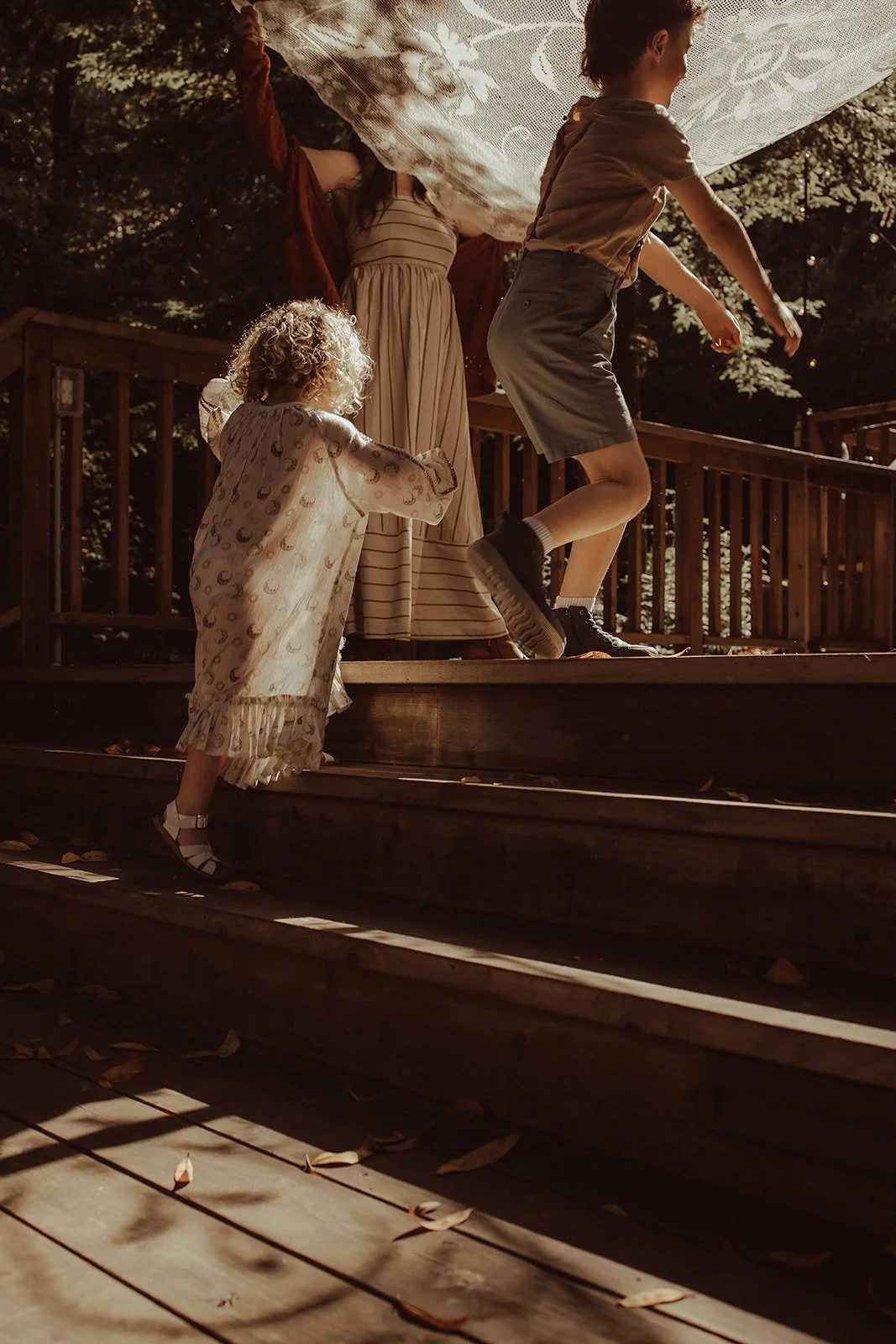 Three children on a wooden outdoor staircase, one girl wearing pajamas, one boy mid-jump, and another girl holding on to the railing, with trees in the background.