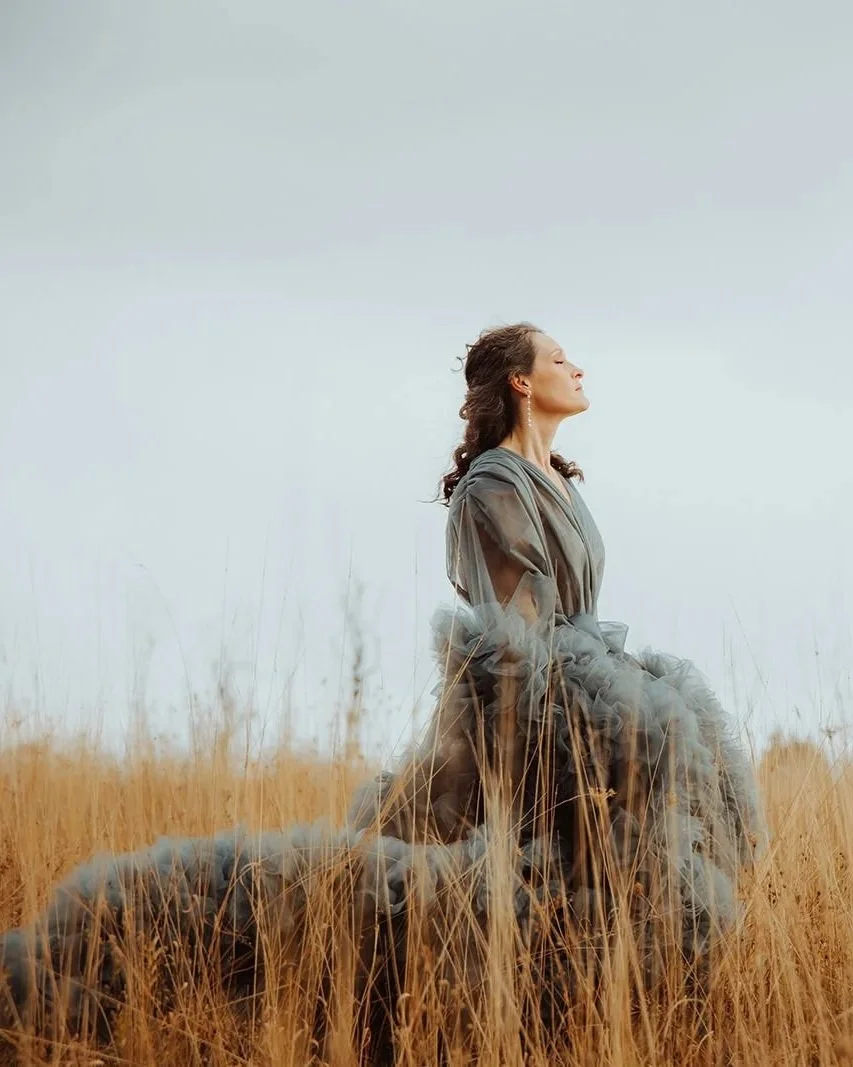 A woman dressed in a flowing gray dress is sitting in a field of tall, dry grass with her eyes closed, appearing to enjoy a moment of peace and tranquility.