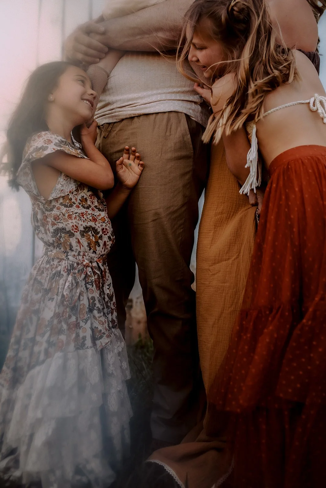 A woman and a young girl are smiling and hugging a man, whose torso is visible, outdoors during daytime.